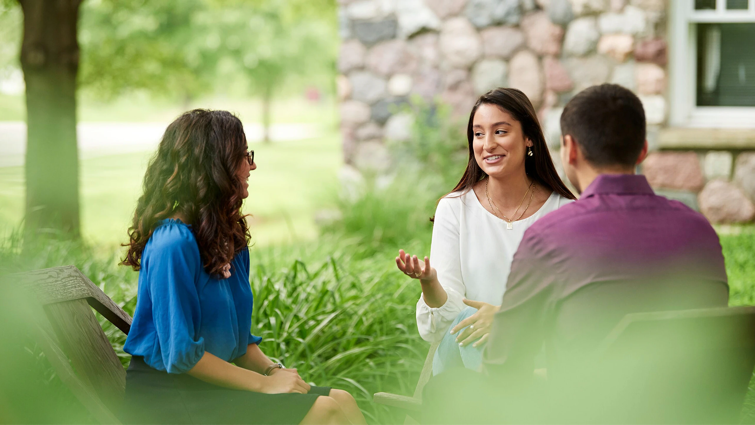 Three people seated in wooden chairs having a conversation outside next to some green foliage and a stone house.