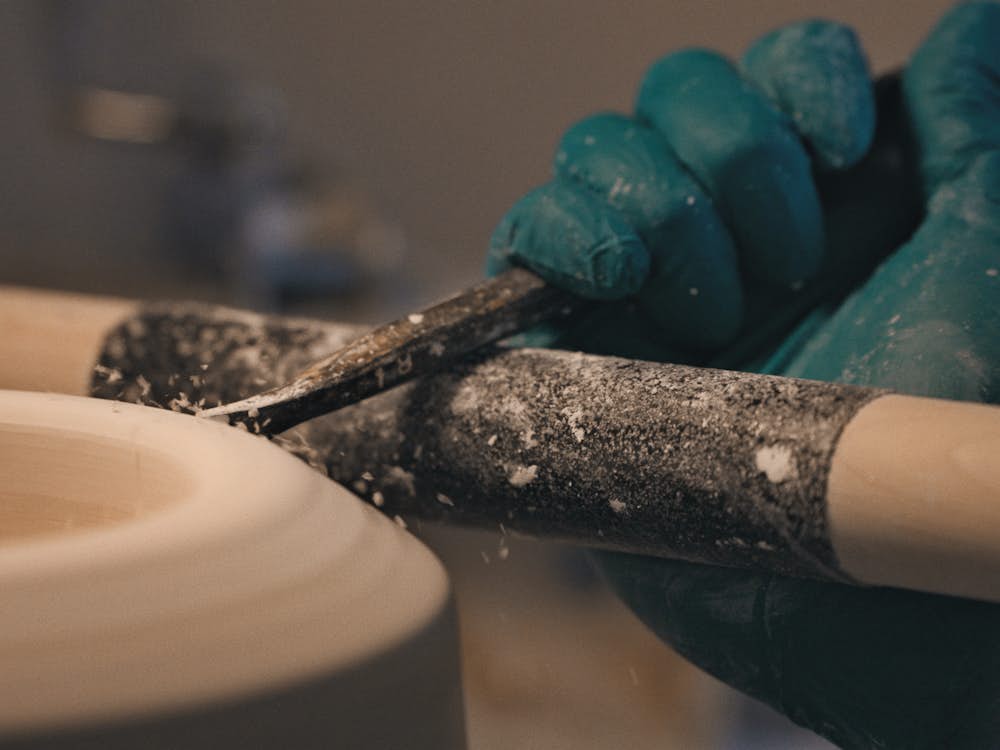 A close-up shot of a gloved hand holding a shaping tool against a piece of greenware, or dried-out clay, spinning on wheel.