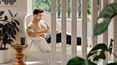 A person with short brown hair, wearing a beige sweater and blue jeans, sitting in a white Knoll Womb Chair and Ottoman while reading next to green potted plants