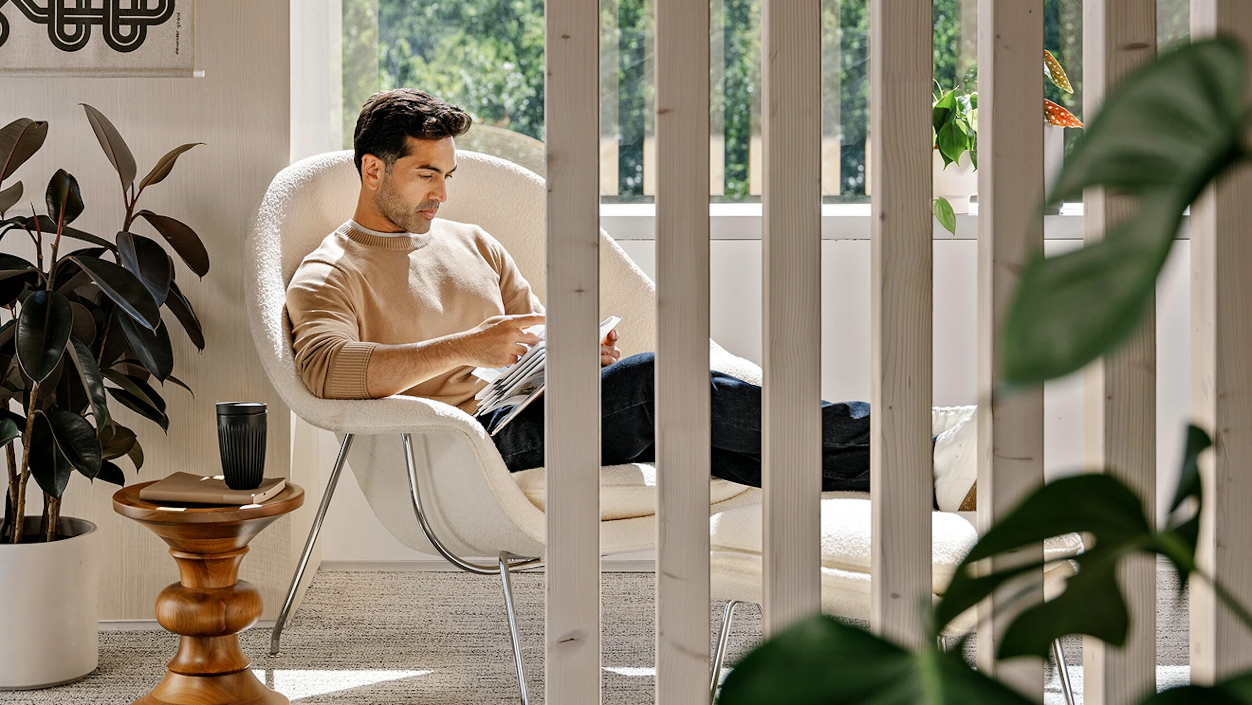 A person with short brown hair, wearing a beige sweater and blue jeans, sitting in a white Knoll Womb Chair and Ottoman while reading next to green potted plants.