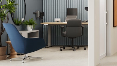 Poltrona azul en primer plano, un escritorio de pie con una computadora portátil y una silla para oficinas en el fondo, paneles de pared con listones y plantas. Un espejo curvo refleja el espacio