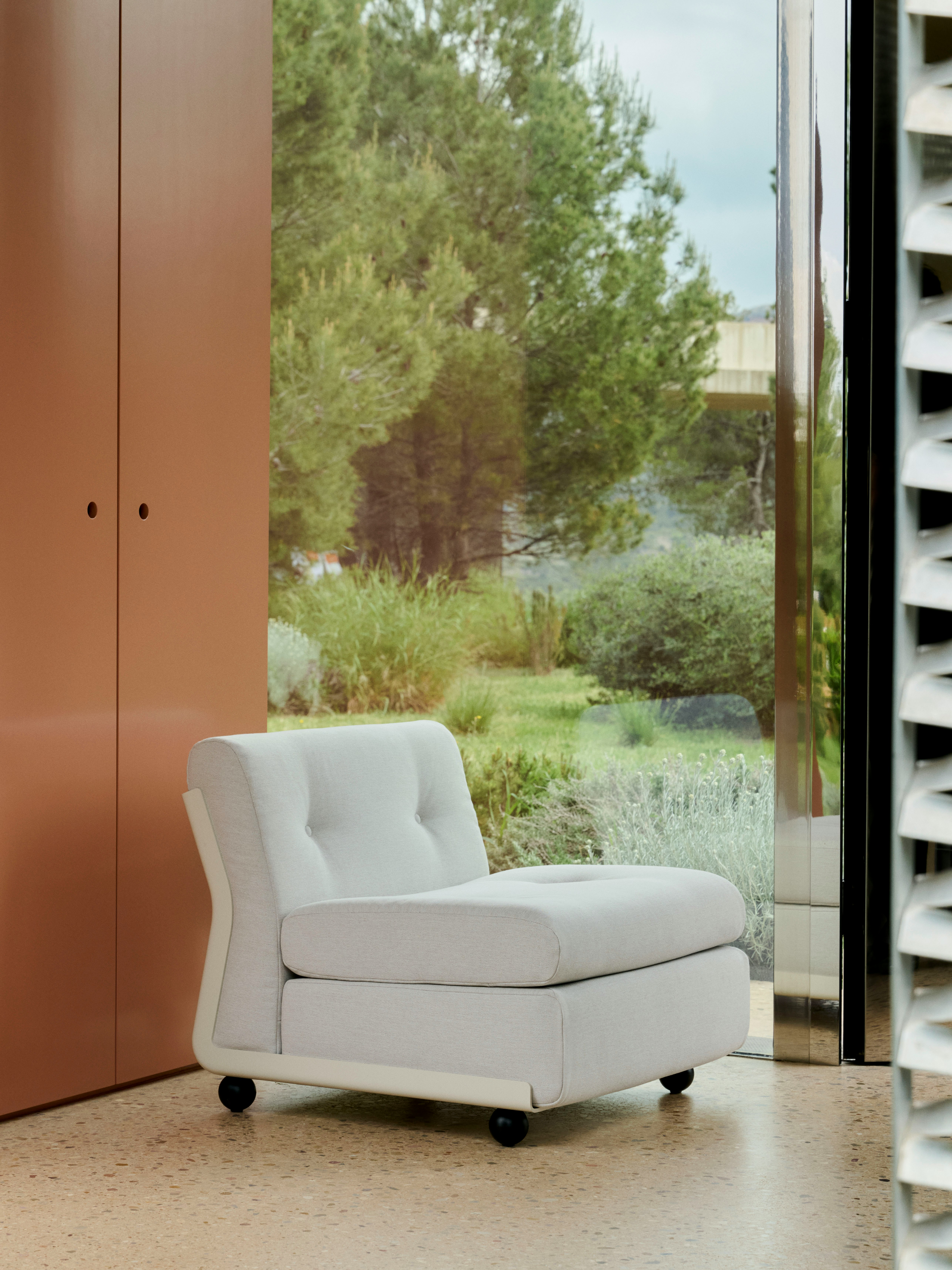 Single-seat white Amanta sofa placed in front of floor-to-ceiling glass with terra-cotta panel backdrop.