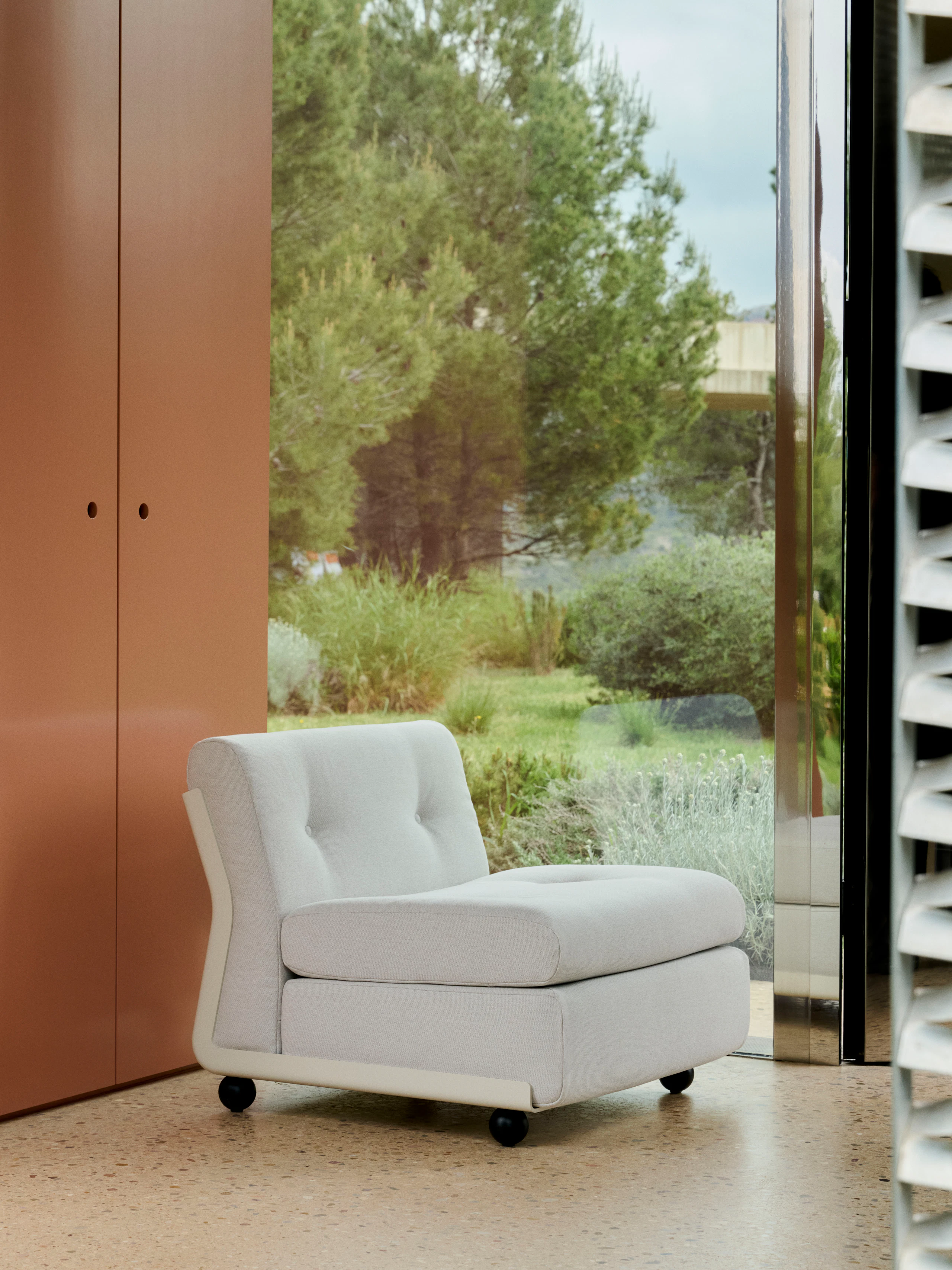 Single-seat white Amanta sofa placed in front of floor-to-ceiling glass with terra-cotta panel backdrop.