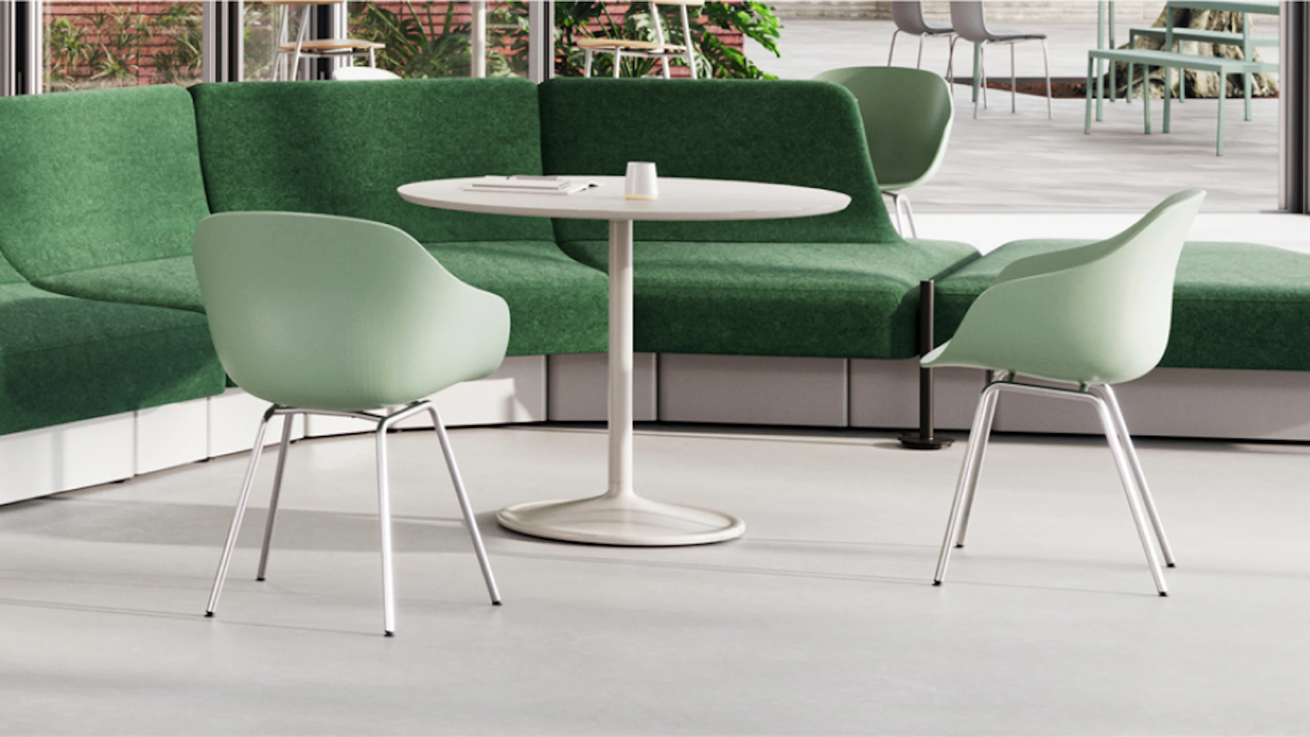 A modern seating area with a round white table, green sectional sofa, and two green chairs on a light floor. Large windows in the background reveal an outdoor space.