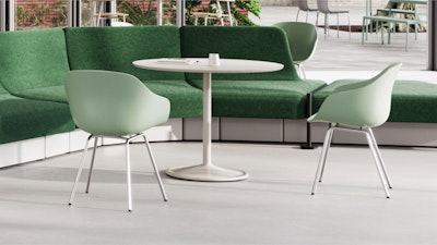 Una moderna zona de estar con una mesa redonda blanca, un sofá seccional verde y dos sillas verdes sobre un suelo claro. Grandes ventanales al fondo dejan ver un espacio exterior.