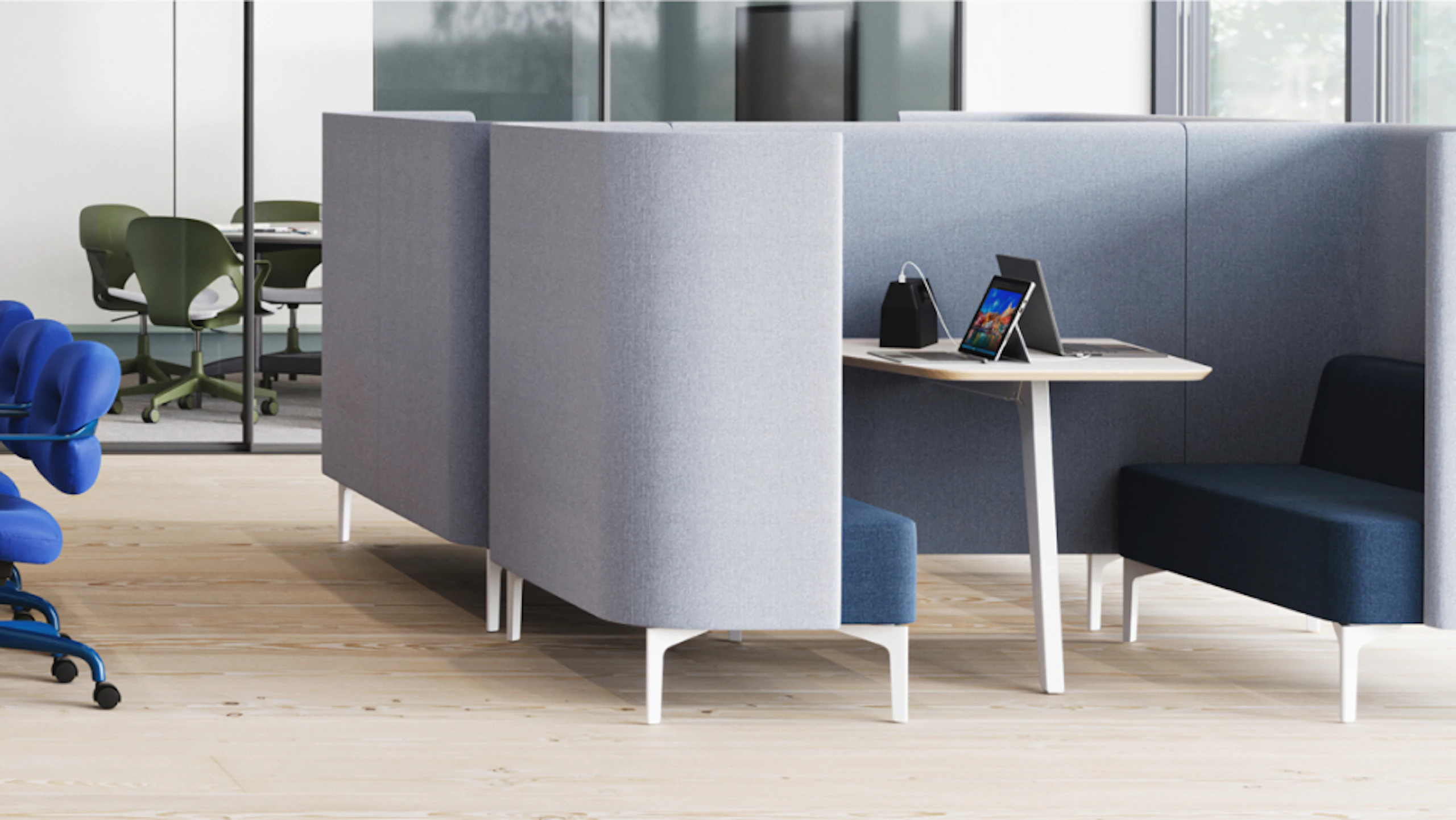 Enclosed booth seating in grey fabric, featuring a small table with a laptop and a dark blue sofa. Rolling office chairs and a table visible in the background.