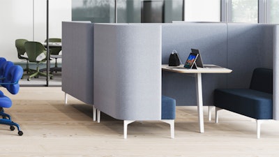 Sillería tipo cabina cerrada en tela gris, con una pequeña mesa con una computadora portátil y un sofá azul oscuro. Sillas para oficinas con ruedas y una mesa visible al fondo.