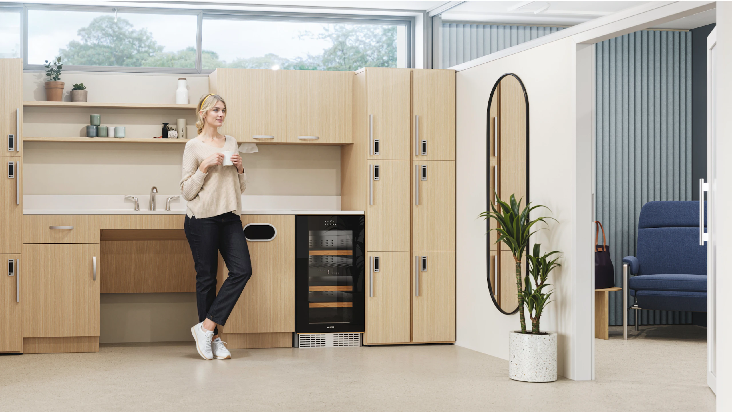 Person takes a break in a modern kitchenette with light wood cabinets, a beverage cooler, and a tall oval mirror. A plant sits in front of the mirror, and a blue armchair is visible in the background.