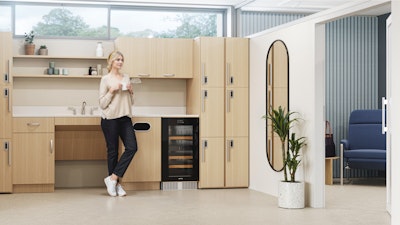 Una persona toma un descanso en una moderna cocina con gabinetes de madera clara, un enfriador de bebidas y un espejo ovalado alto. Delante del espejo hay una planta y al fondo se ve una poltrona azul.