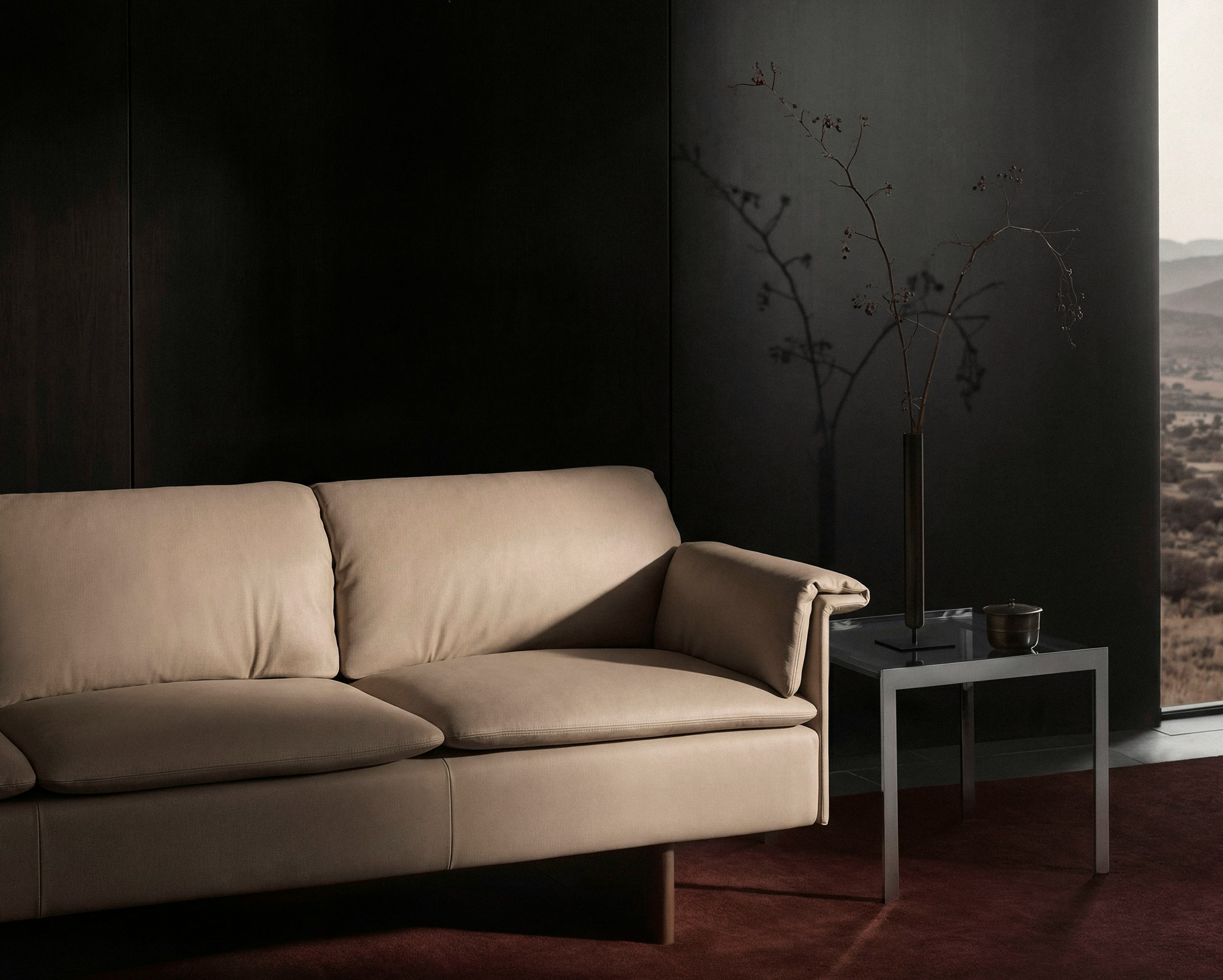 Mantle Sofa designed by Ward Bennett upholstered with Bristol Leather in Champagne beside an H Frame side table, viewed at an angle against a black wall on a burgundy rug.