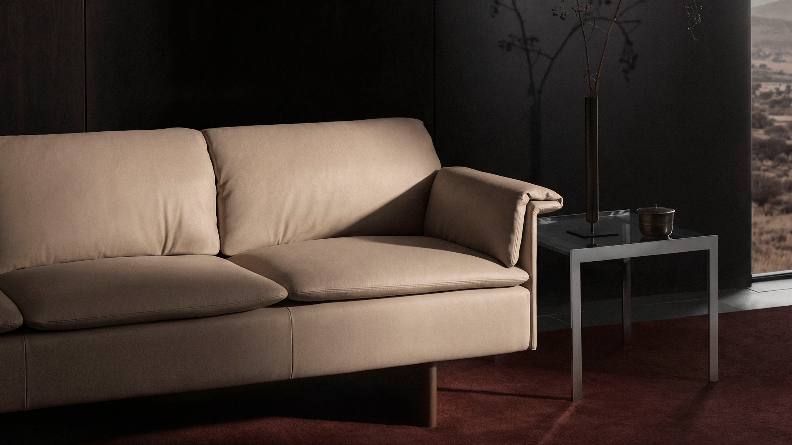 Mantle Sofa designed by Ward Bennett upholstered with Bristol Leather in Champagne beside an H Frame side table, viewed at an angle against a black wall on a burgundy rug.