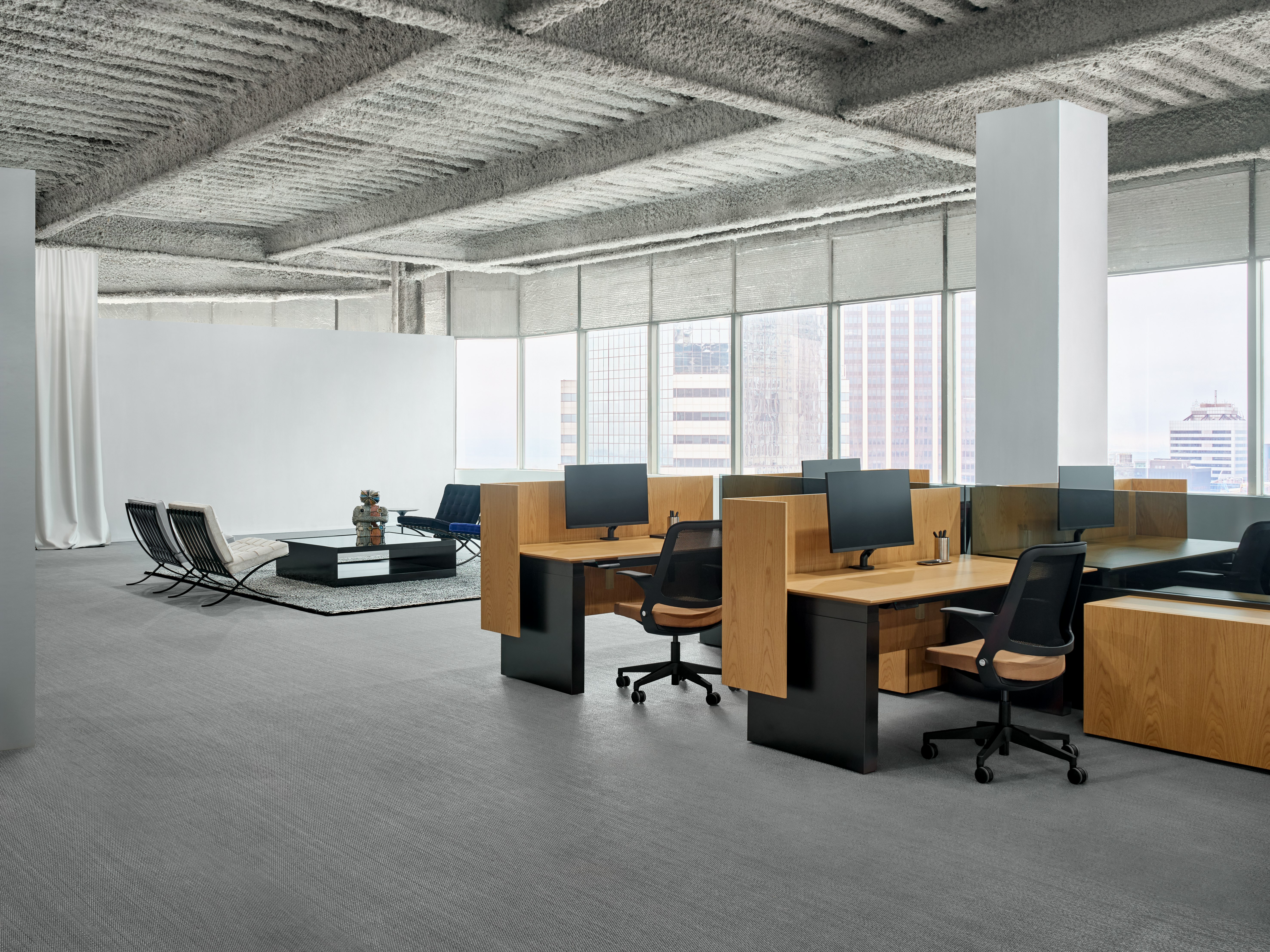 A group of workstations are grouped together and each station has a height adjustable table, a work chair and personal storage. In the background are some lounge chairs around a coffee table.