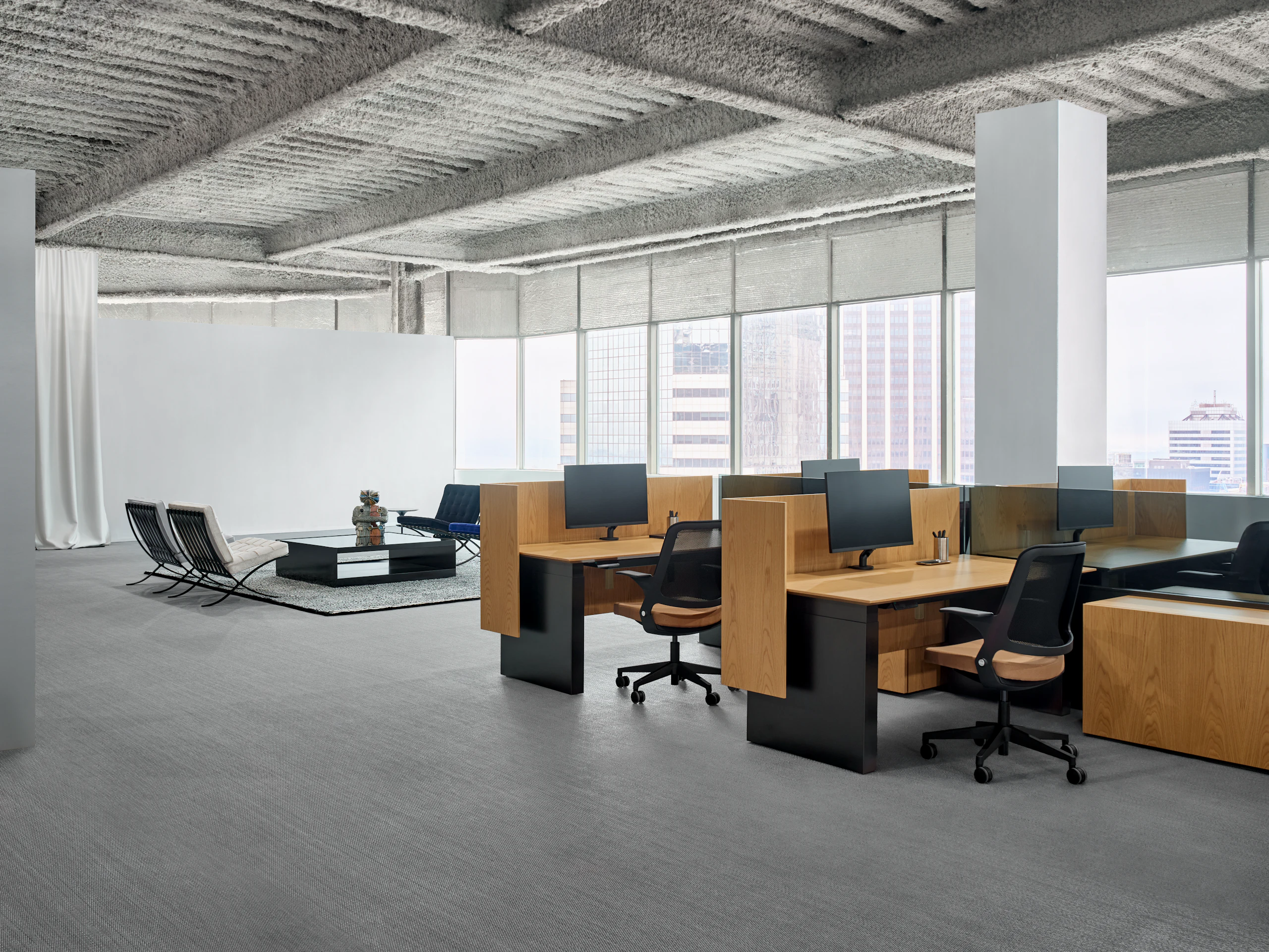 A group of workstations are grouped together and each station has a height adjustable table, a work chair and personal storage. In the background are some lounge chairs around a coffee table.