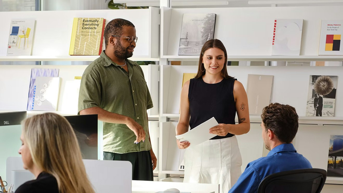 Several people having a conversation around an individual workstation furnished with Herman Miller Aeron Chairs and sit-to-stand desks.
