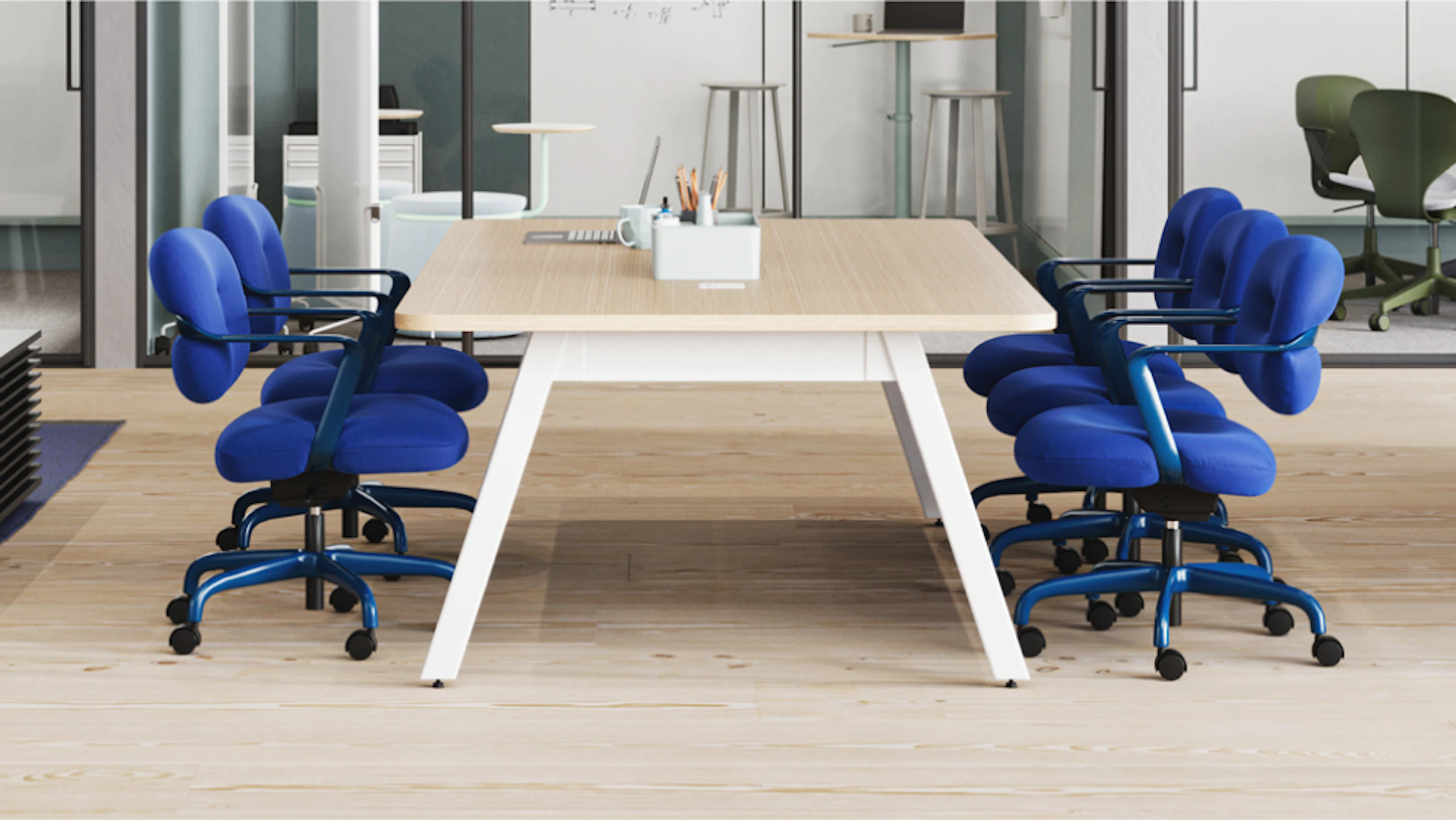 Light wood table surrounded by bright blue rolling chairs. Pencil cup and other items on the table. Background shows other workspaces and stools.