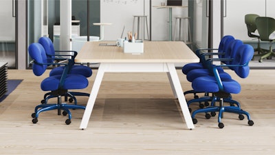 Mesa de madera clara rodeada de sillas con ruedas de color azul brillante. Portalápices y otros objetos sobre la mesa. El fondo muestra otros espacios de trabajo y taburetes.