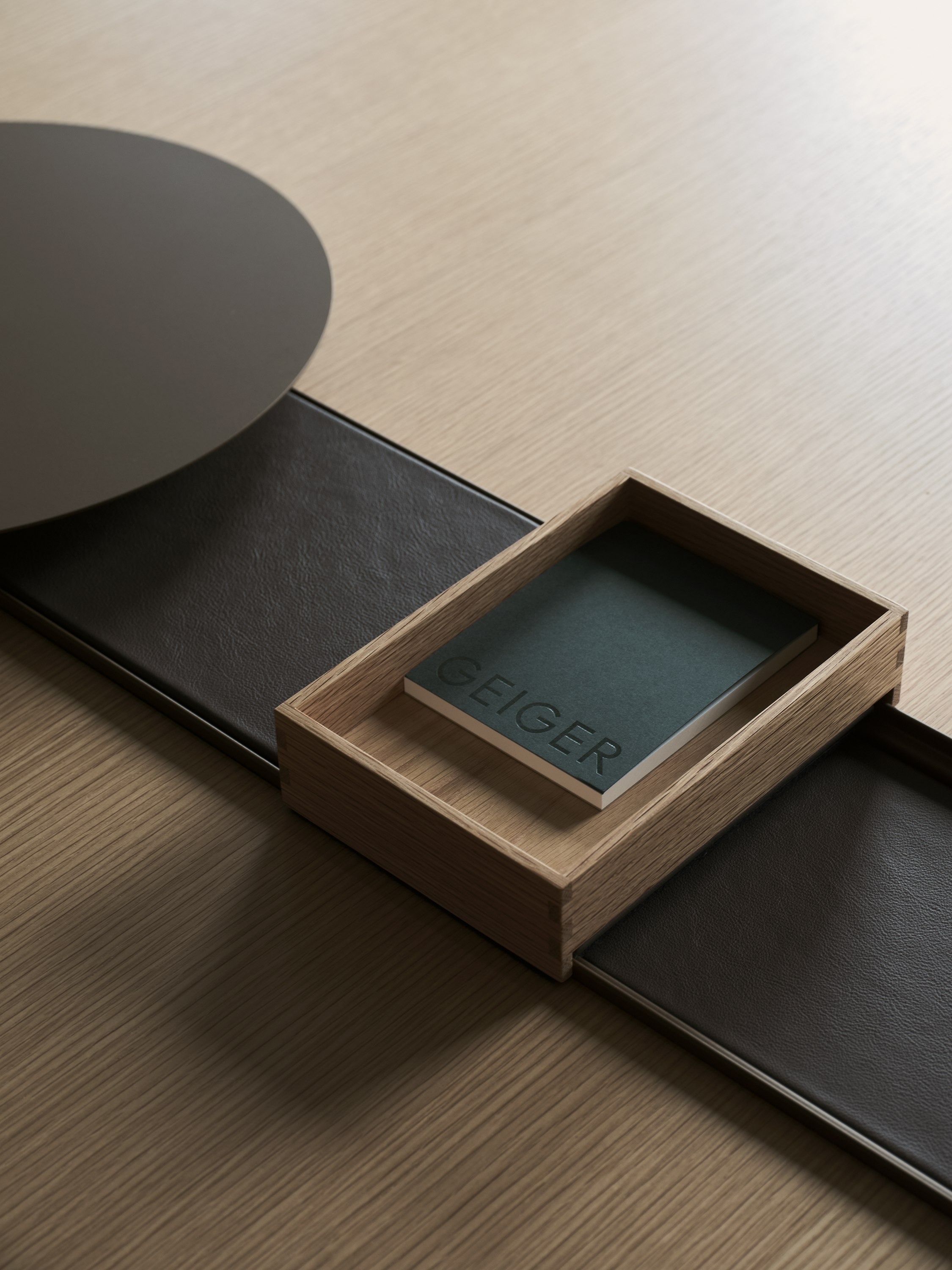 Detail view of a closed Ascent Conference Table power trough with leather covers, an AV platform and an accessory tray containing a Geiger notebook, with a Natural Rift Cut Oak tabletop, viewed from above at an angle.