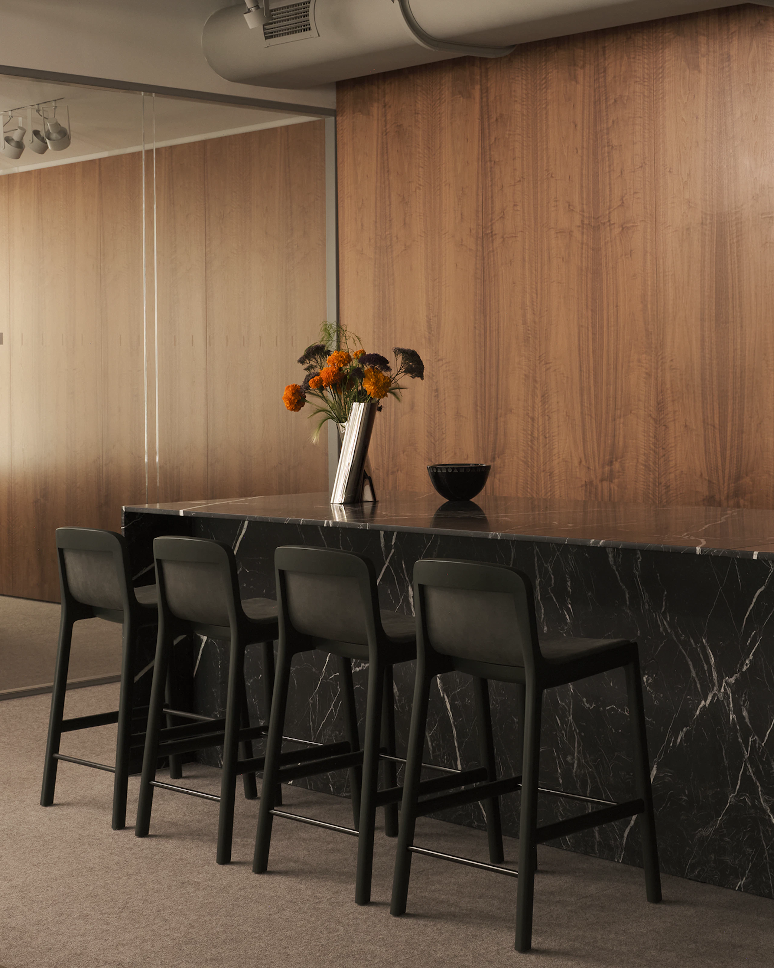 A custom Geiger reception desk in Nero Marquina Marble with four counter height Lijn Stools in Ivy and a Clamshell Chair against a veneer wall in the Geiger Chicago showroom, viewed at an angle.