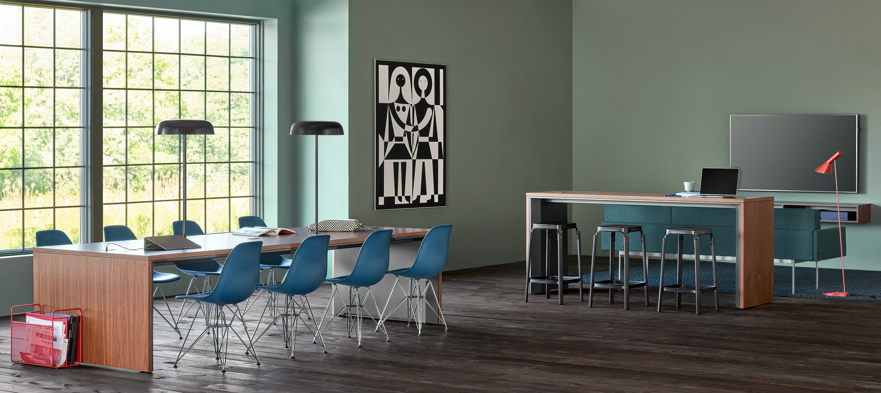 A community space with a Headway communal table surrounded by Eames Plastic side chairs and another nearby communal table behind a couch facing a TV.
