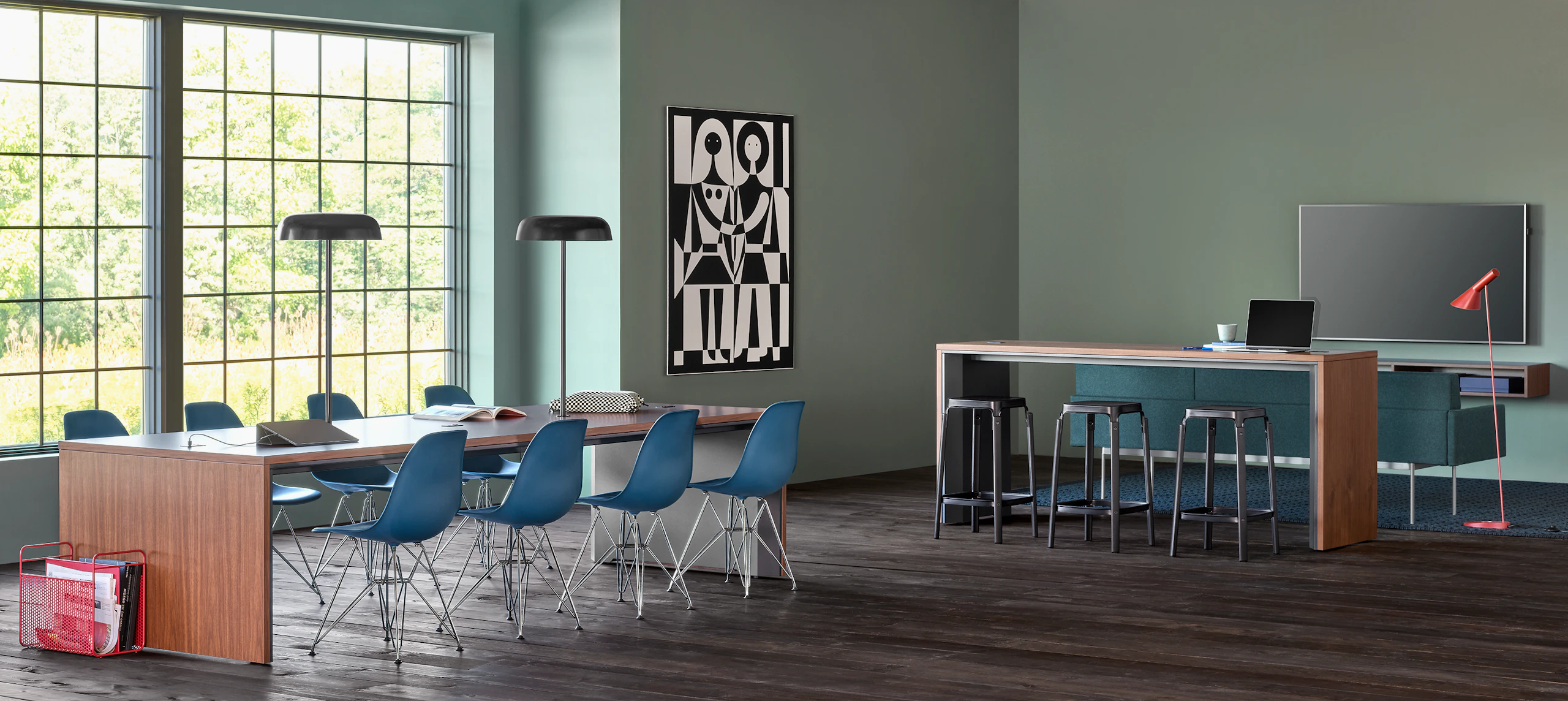 A community space with a Headway communal table surrounded by Eames Plastic side chairs and another nearby communal table behind a couch facing a TV.