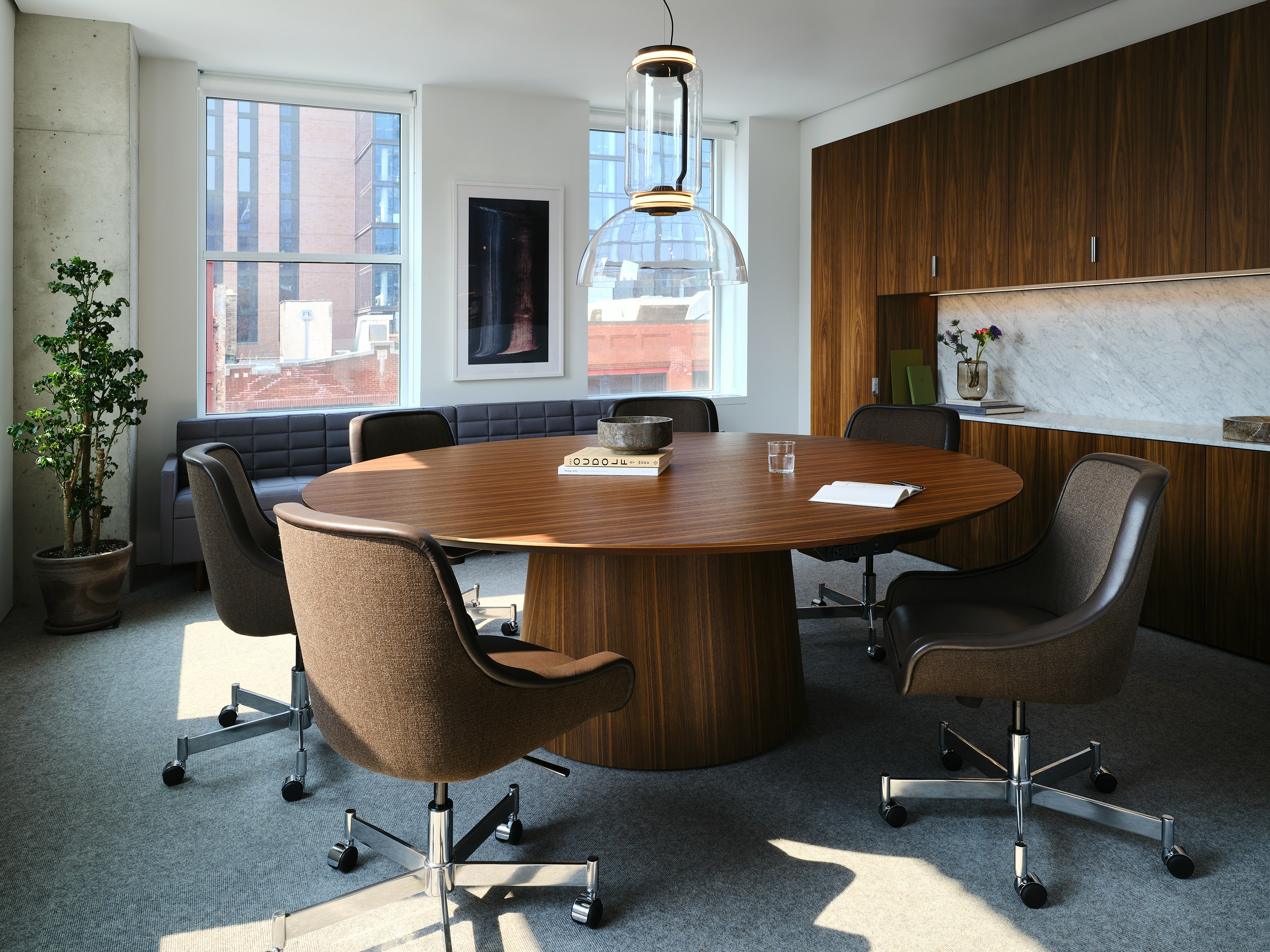 JD Meeting Table in Natural Quarter Cut Walnut with round top and tapered base shown at an angle surrounded by Bumper Chairs in front of a Geiger One workwall.