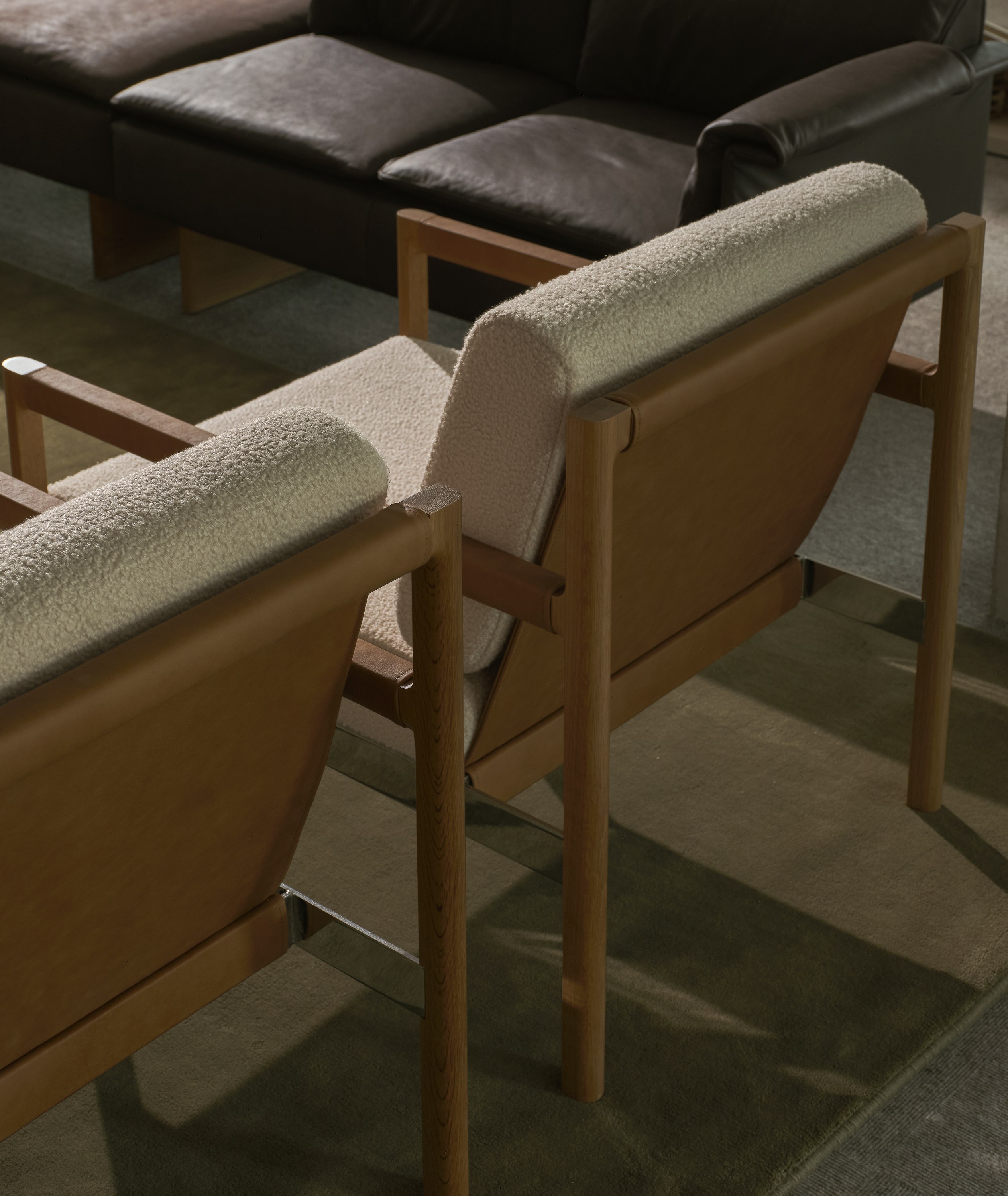 Detail view of the backs of two Brabo Club Chairs with Natural Oak and chrome frames and upholstered with Maharam Firth in Limewash and a Mantle Sectional Sofa in the background, viewed from above at an angle.