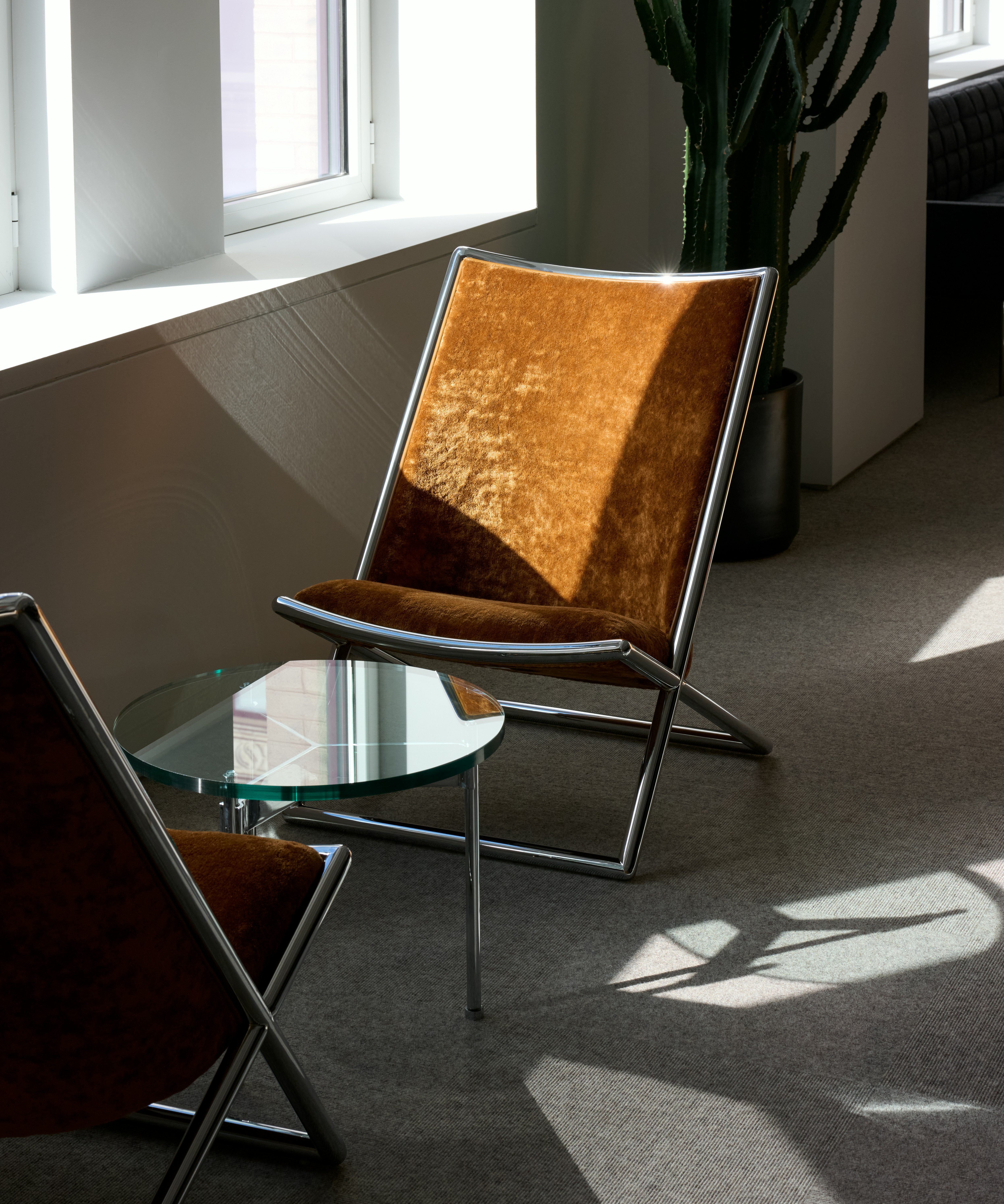 Two Scissor Chairs with burnt orange upholstery and chrome frames with a round Claw Table with a glass top and chrome base, viewed at an angle in a showroom setting.