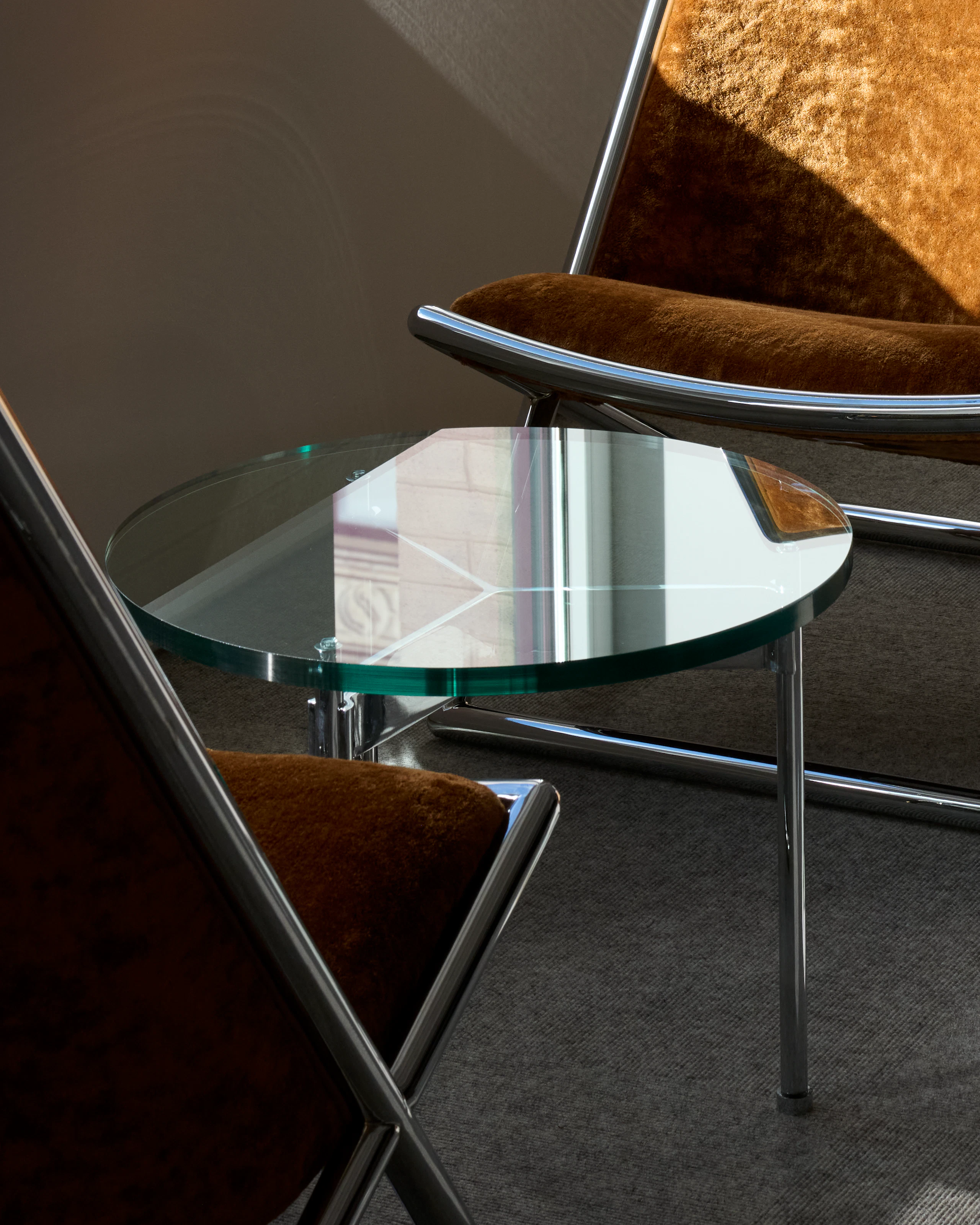Two Scissor Chairs with burnt orange upholstery and chrome frames with a round Claw Table with a glass top and chrome base, viewed at an angle in a showroom setting.