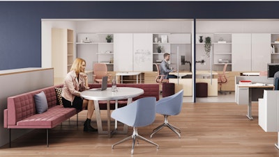 Un espacio de trabajo moderno con piso de madera, con una persona sentada en una mesa redonda, junto con una persona que trabaja en un escritorio detrás de un vidrio y muebles contemporáneos, que incluyen sillas rojas y azules.