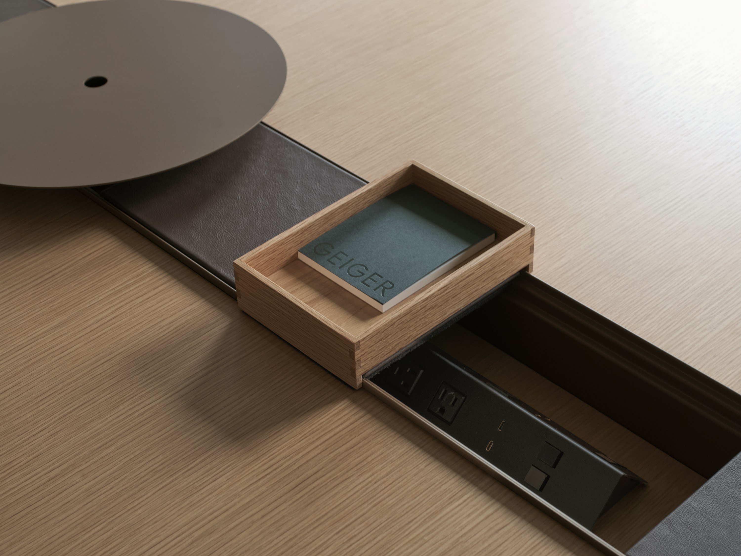 Detail view of an open Ascent Conference Table power trough with leather covers, an AV platform and an accessory tray containing a Geiger notebook, with a Natural Rift Cut Oak tabletop, viewed from above at an angle.