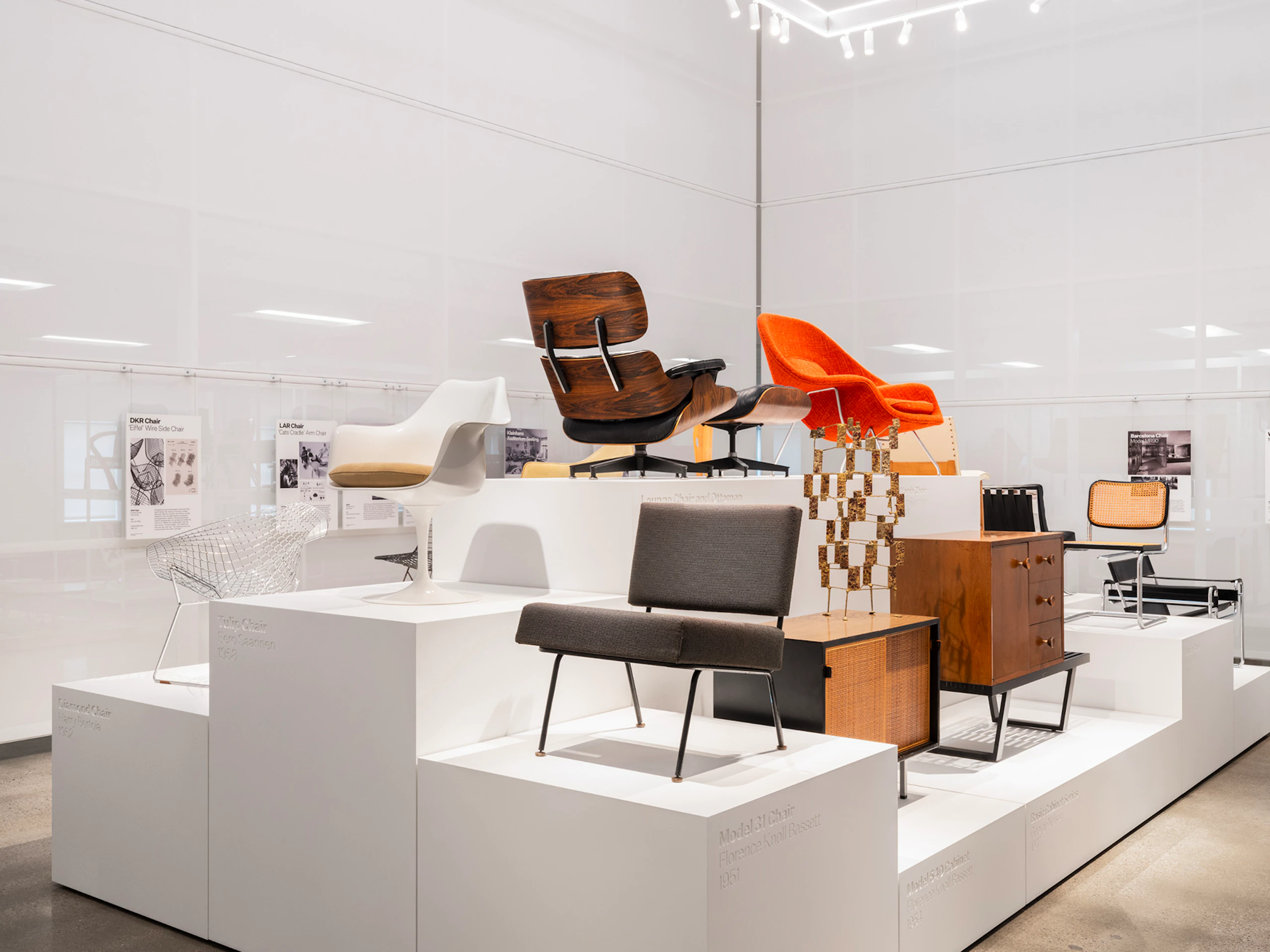Classic pieces of furniture from the MillerKnoll collective, such as an orange Knoll Womb Chair and black Knoll Barcelona Chair, arranged on white pedestals at the MillerKnoll Archives