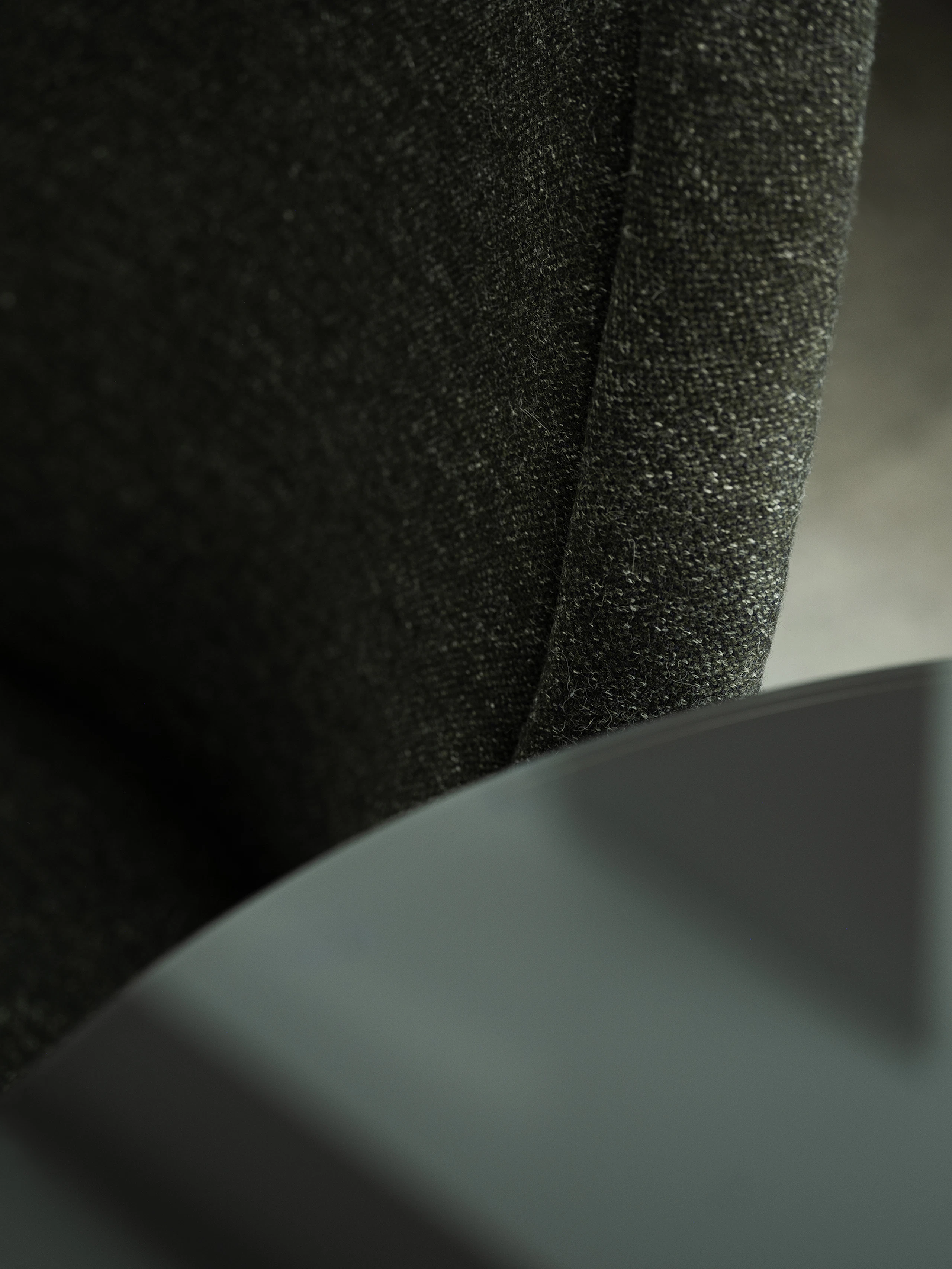 Detail view of a Bumper Conference Chair with low arms designed by Ward Bennet, upholstered with Bellano in Black Green, viewed at an angle next to a glass tabletop.