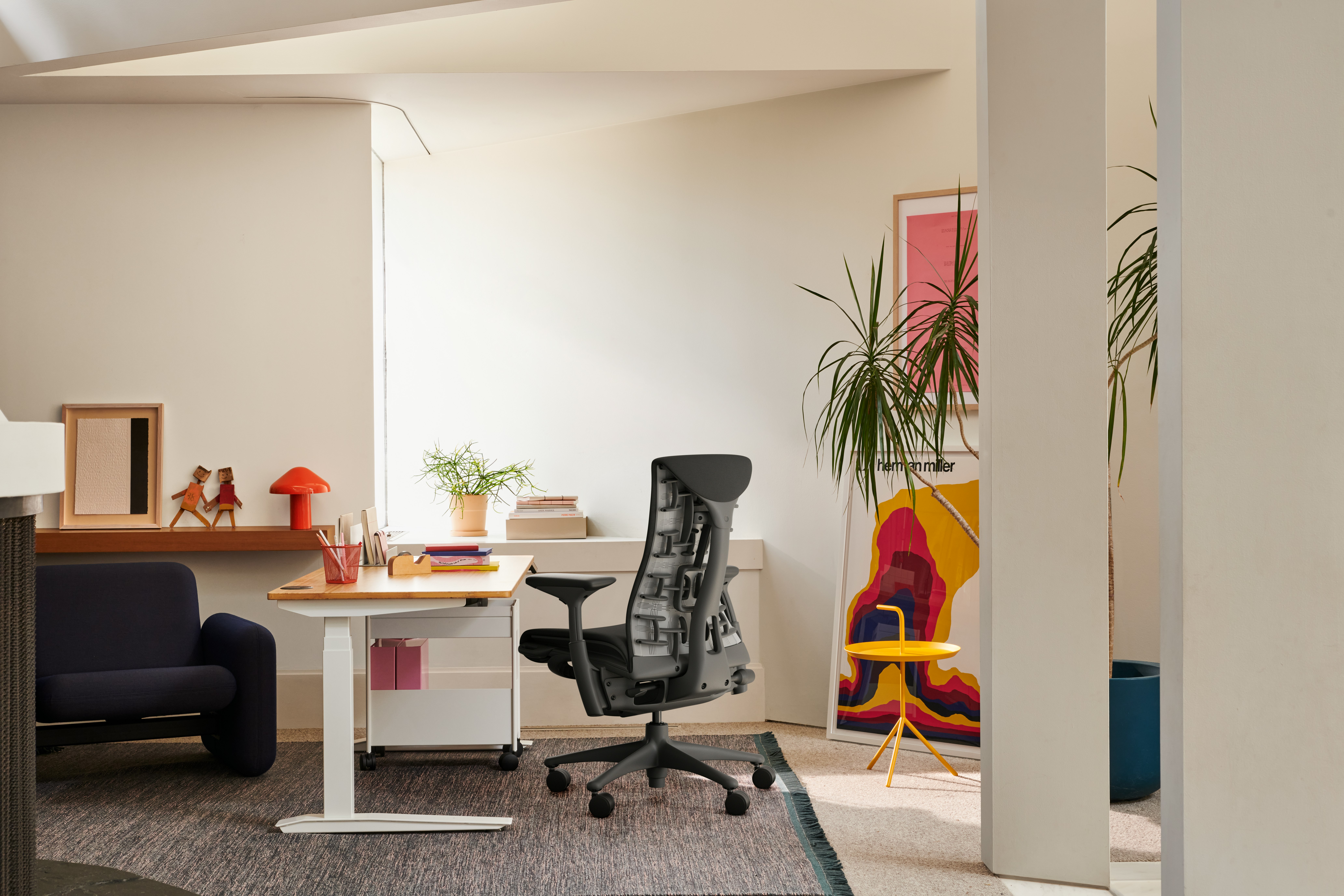 Side view of a bright office featuring a black graphite Herman Miller Embody Office Chair, Jarvis Bamboo Sit-Stand Desk, Wilkes Modular Armchair, HAY Pao Portable Lamp and OE1 Storage Trolley.