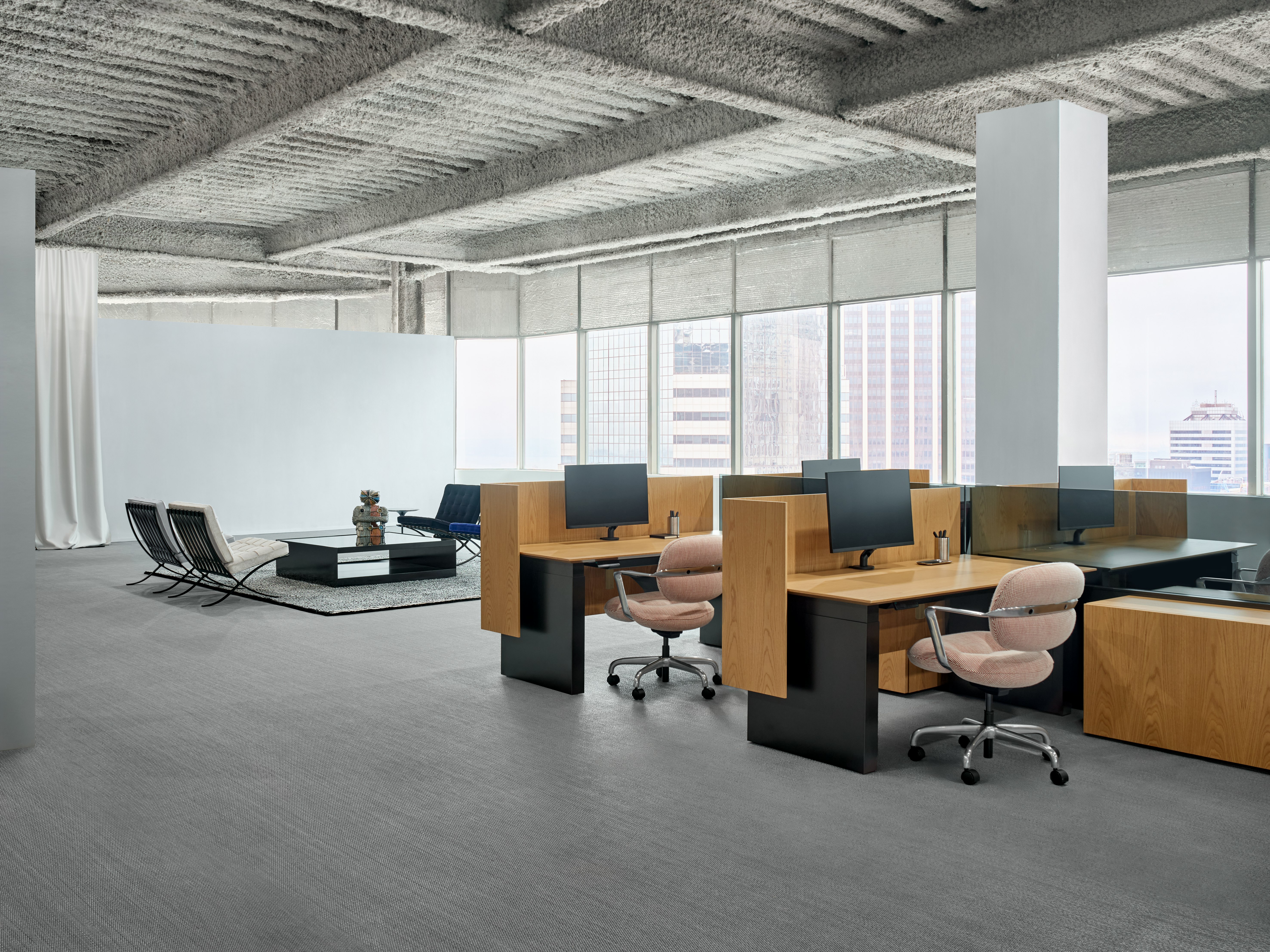 A group of workstations is in an open office in front of a large wall of windows. Behind the workstations is a lounge seating area with a square coffee table between the chairs.