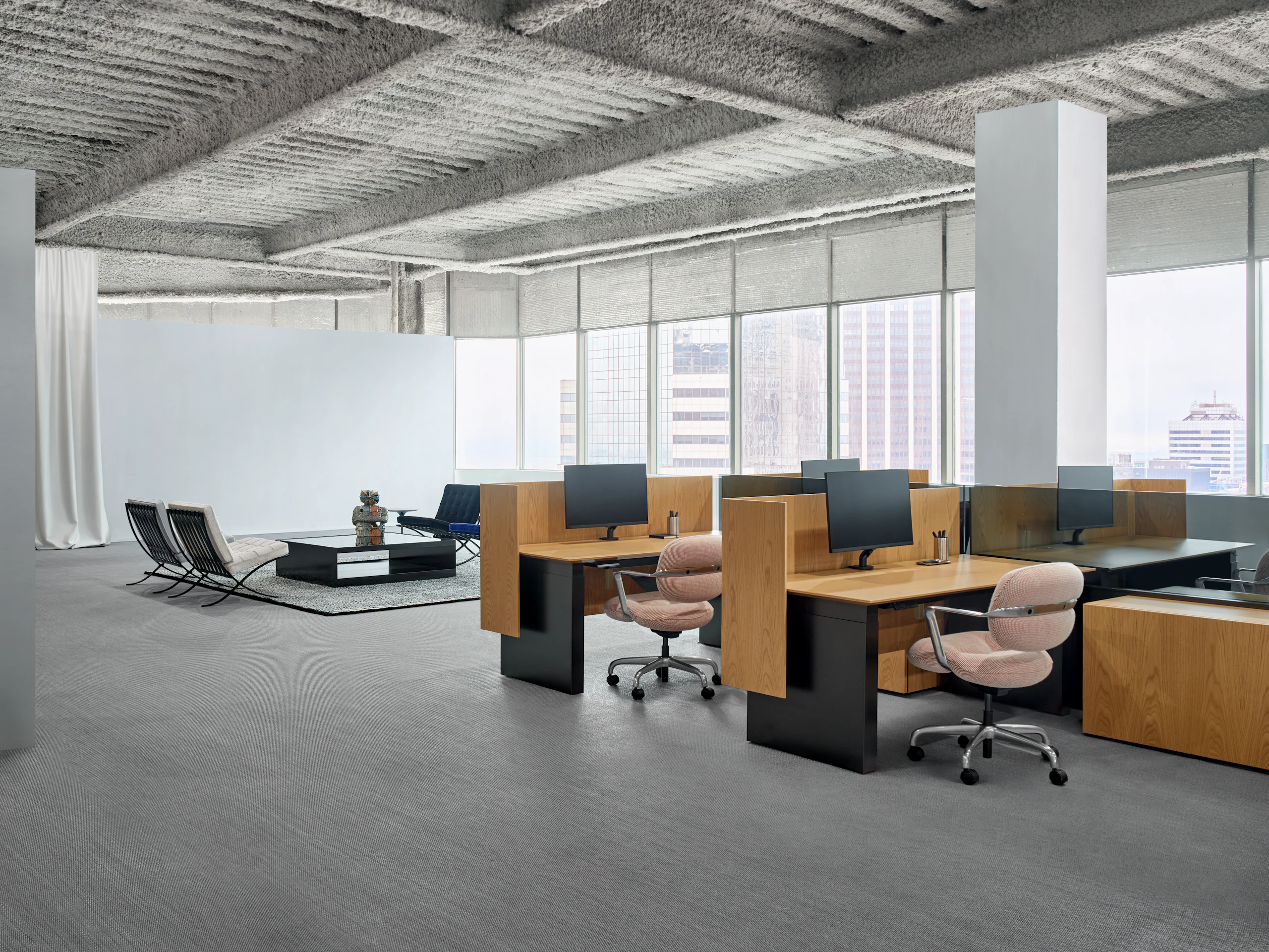 A group of workstations is in an open office in front of a large wall of windows. Behind the workstations is a lounge seating area with a square coffee table between the chairs.