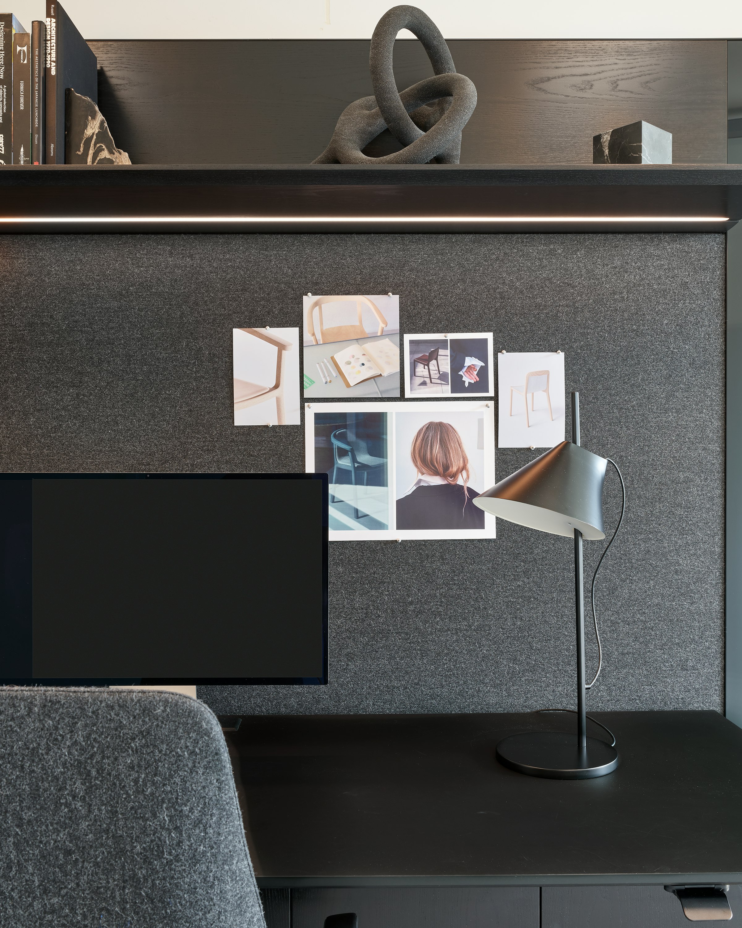Detail view of a Geiger One Casegoods work surface, tackboard, and overhead shelving with integrated lighting in Ebonized Flat Cut Ash and a grey textile, viewed from the front in a private office setting. 