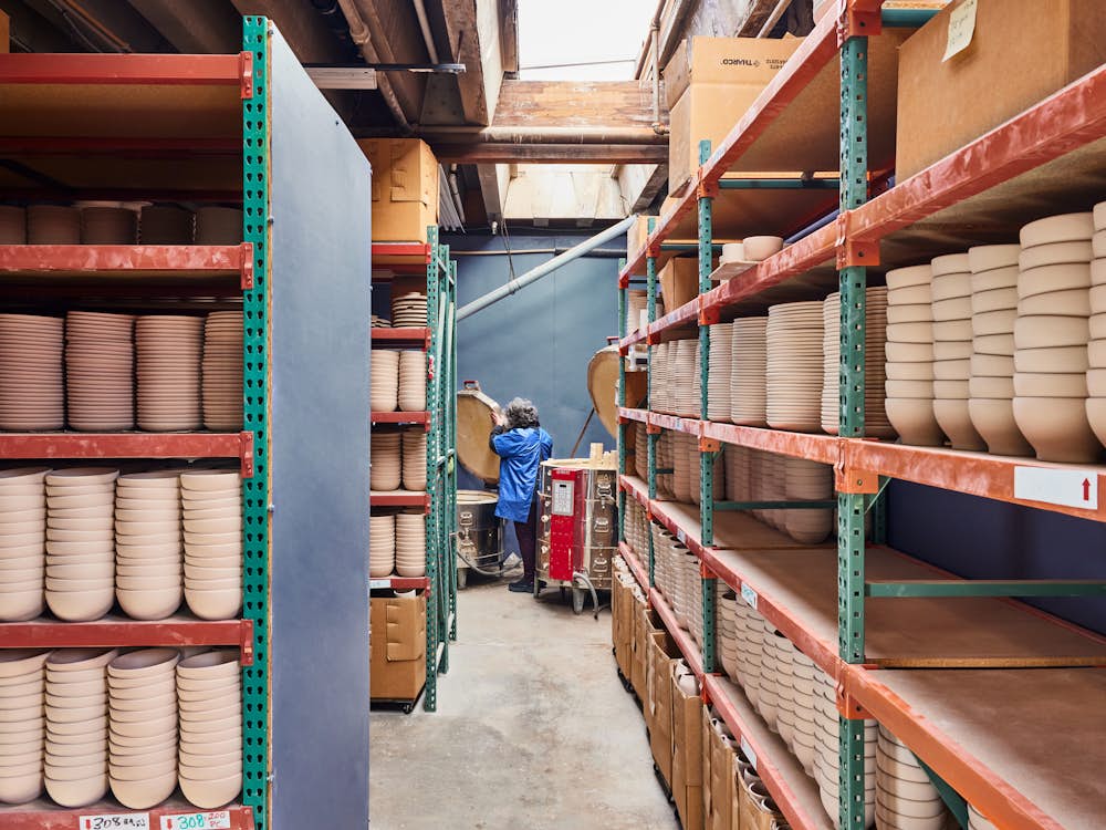 Stacks of unglazed bowls and plates sit on red and green shelving with a glazer in the background readying pieces.