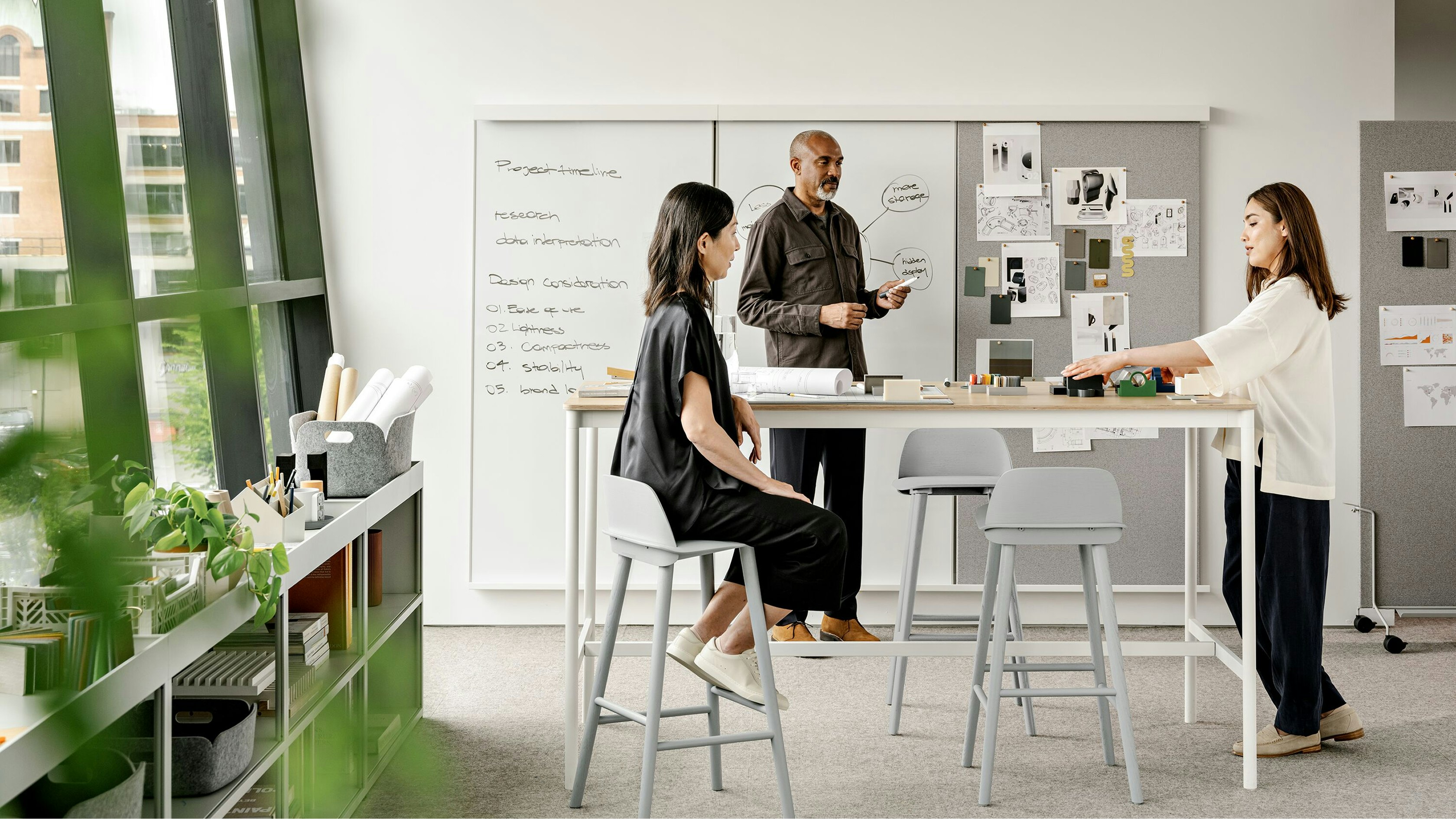 A man and two women working in front of a whiteboard around a tall counter height table with stools.