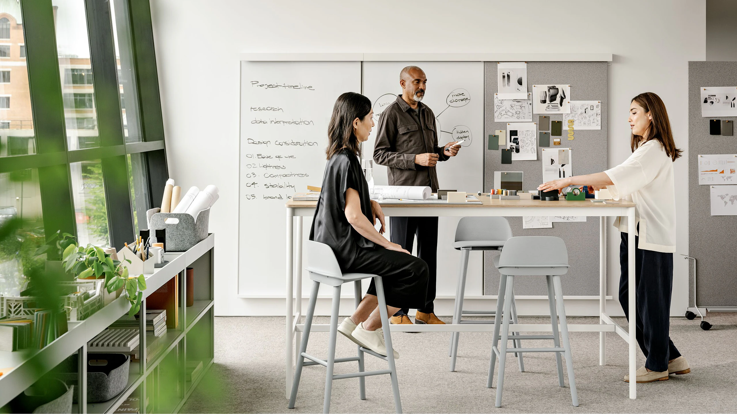 A man and two women working in front of a whiteboard around a tall counter height table with stools.