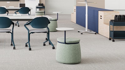 Aula moderna con una mesa larga blanca con sillas color verde azulado y taburetes móviles únicos con pequeños escritorios adjuntos. Al fondo se encuentran un espacio de almacenamiento modular y un escritorio para trabajar de pie.