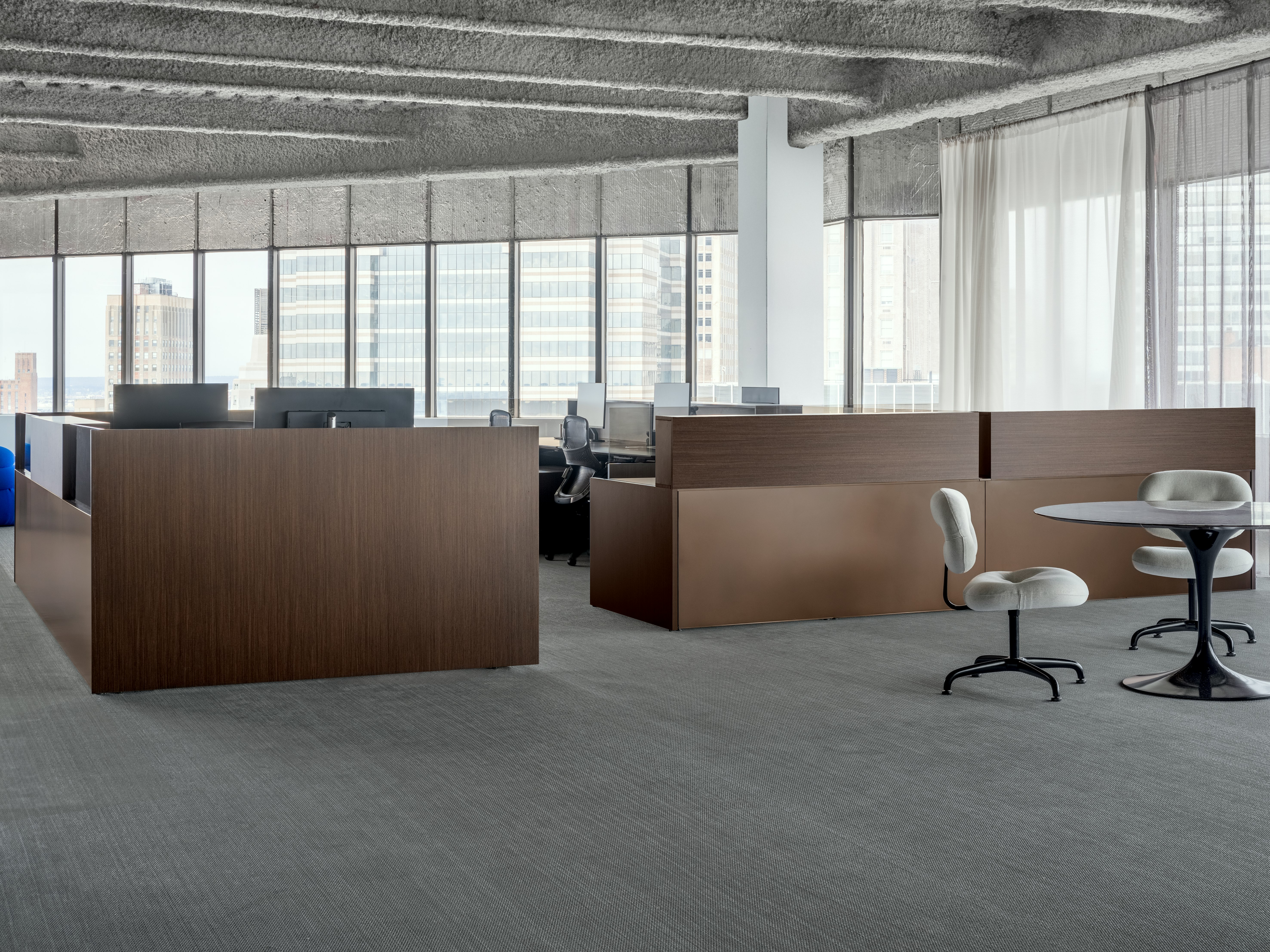 A group of workstations are positioned in front of a large wall of windows. To the side of the workstations is a small meeting table and side chairs.