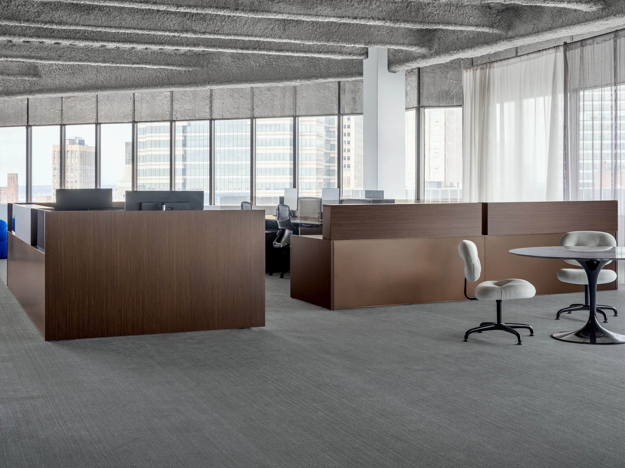A group of workstations are positioned in front of a large wall of windows. To the side of the workstations is a small meeting table and side chairs.