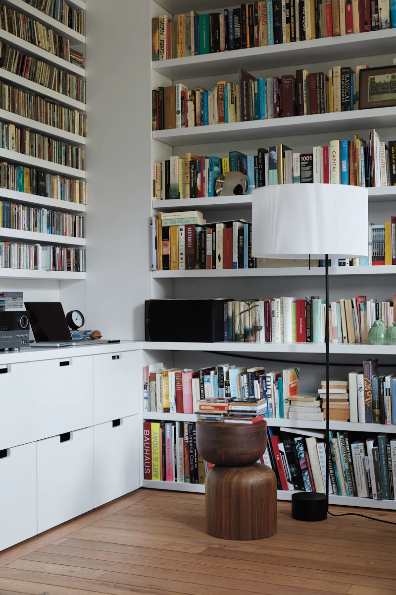Extensive bookshelf wall with white and wooden shelving filled with books; round wooden stool and lamp