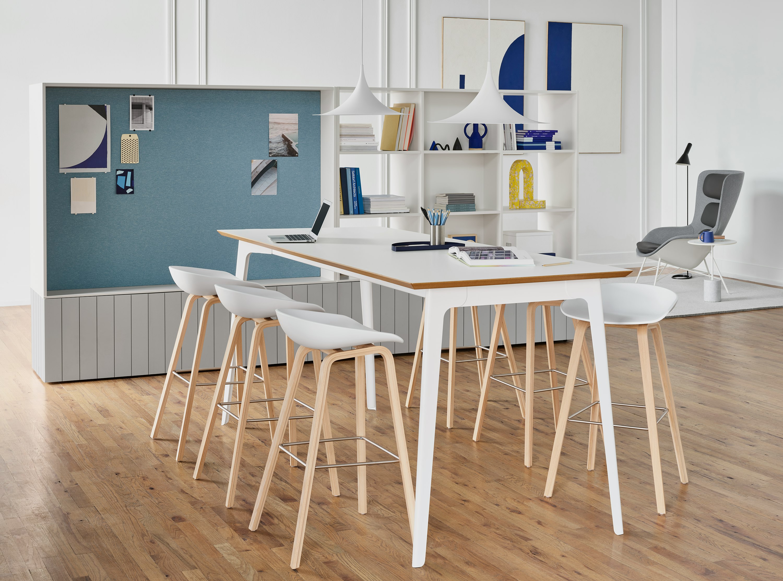 Six About A Stool barstools in white with a wood base around a white Fold Bar Height Table. The scene is set in front of a white shelving unit and blue display tackboards.