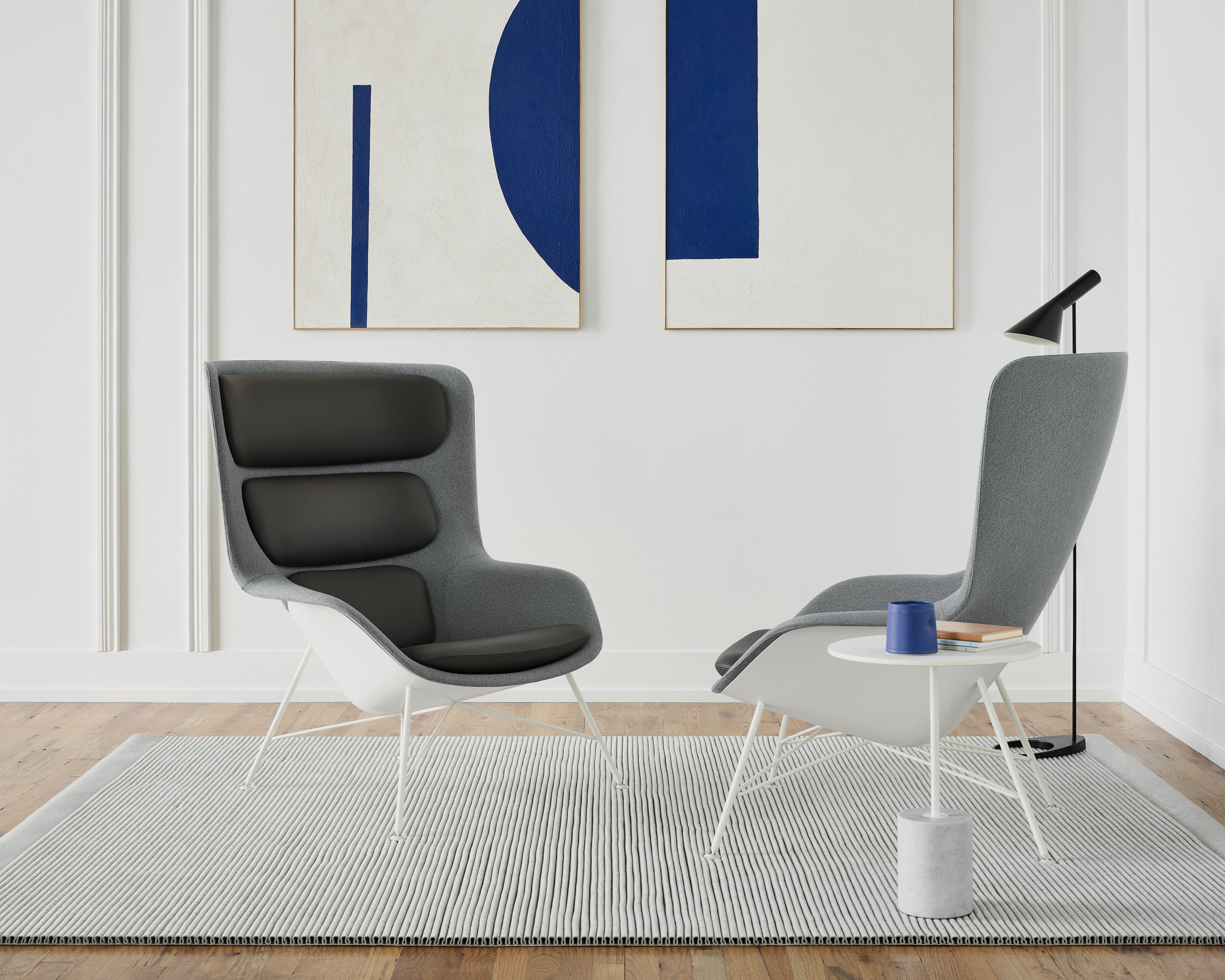Two grey Straid Lounge Chairs next to a white Jey table and a black AJ Floor Lamp. The scene is set on a light grey rug in front of a wall with two picture frames.