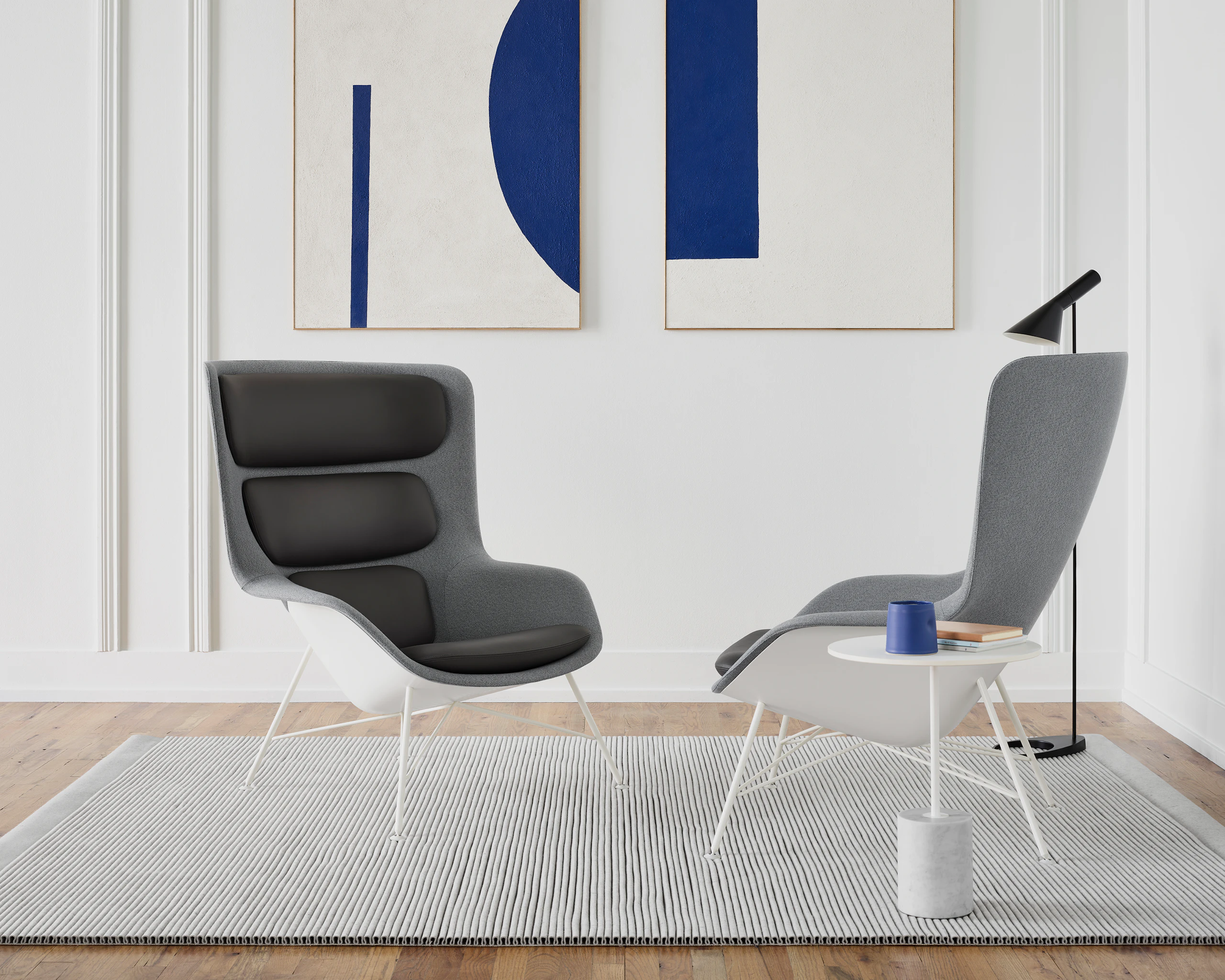 Two grey Straid Lounge Chairs next to a white Jey table and a black AJ Floor Lamp. The scene is set on a light grey rug in front of a wall with two picture frames.