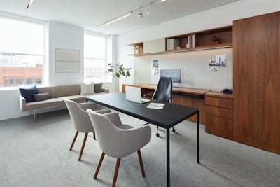 A black desk with two grey guest chairs in front of it sit in a spacious private office with dark wood shelves and storage in the background.