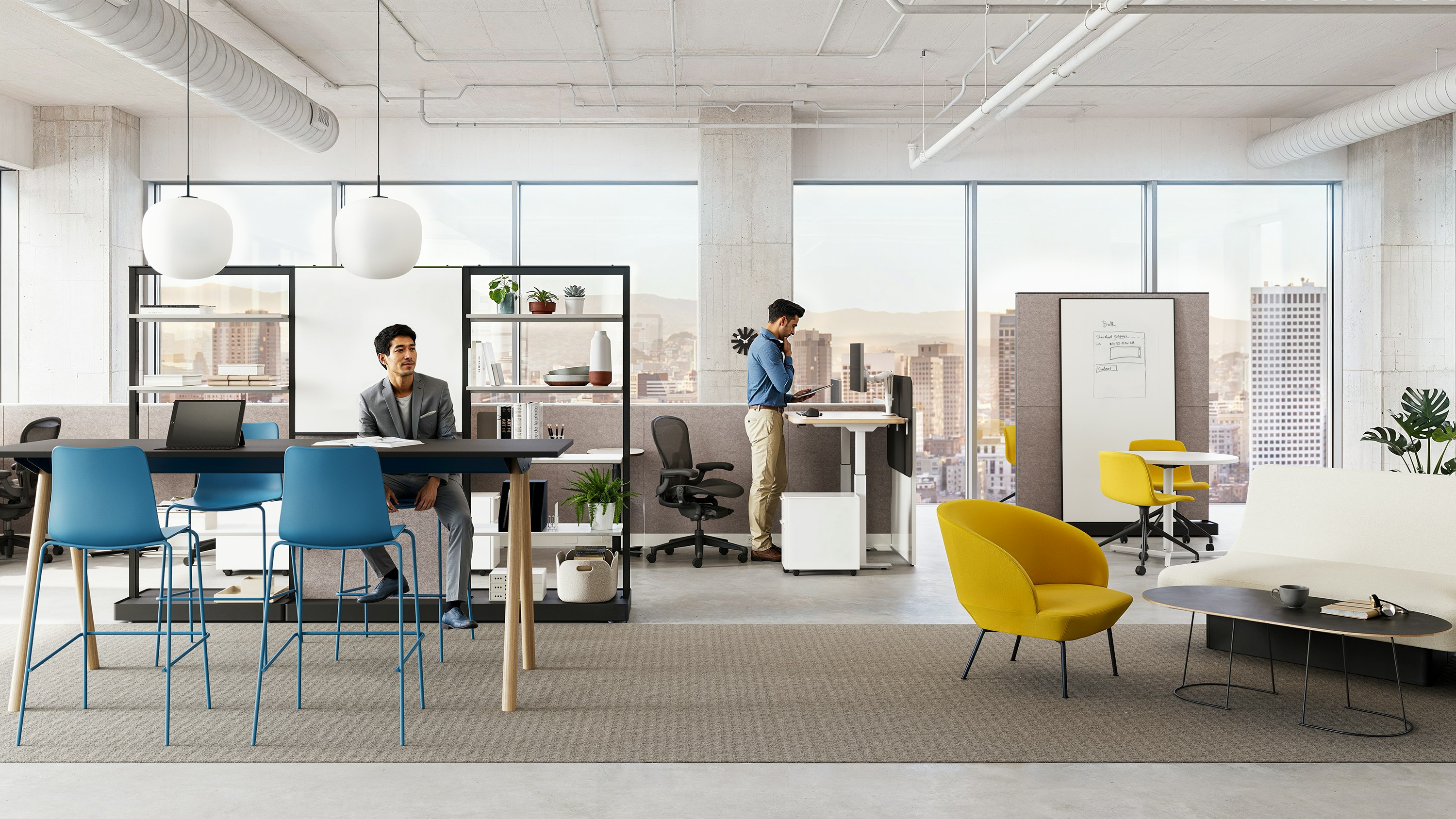 Two people in a workplace setting furnished with a Morse Table System, black Aeron Chairs, blue Polly counter-height stools, and items from the OE1 Workplace Collection.