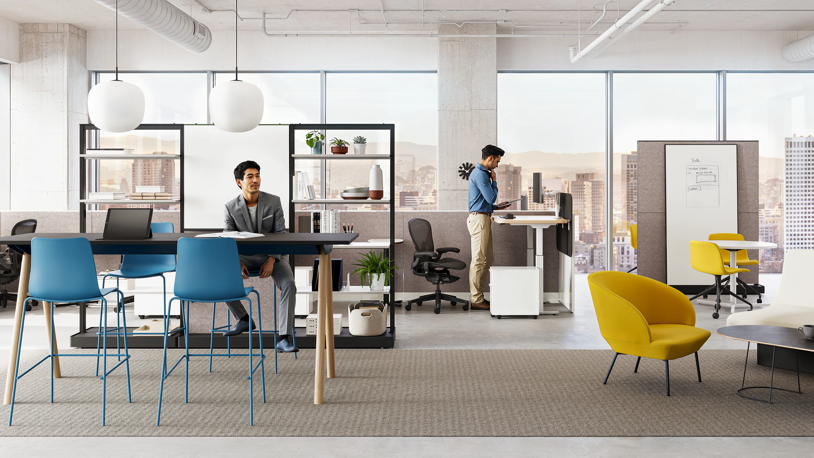 Two people in a workplace setting furnished with a Morse Table System, black Aeron Chairs, blue Polly counter-height stools, and items from the OE1 Workplace Collection.