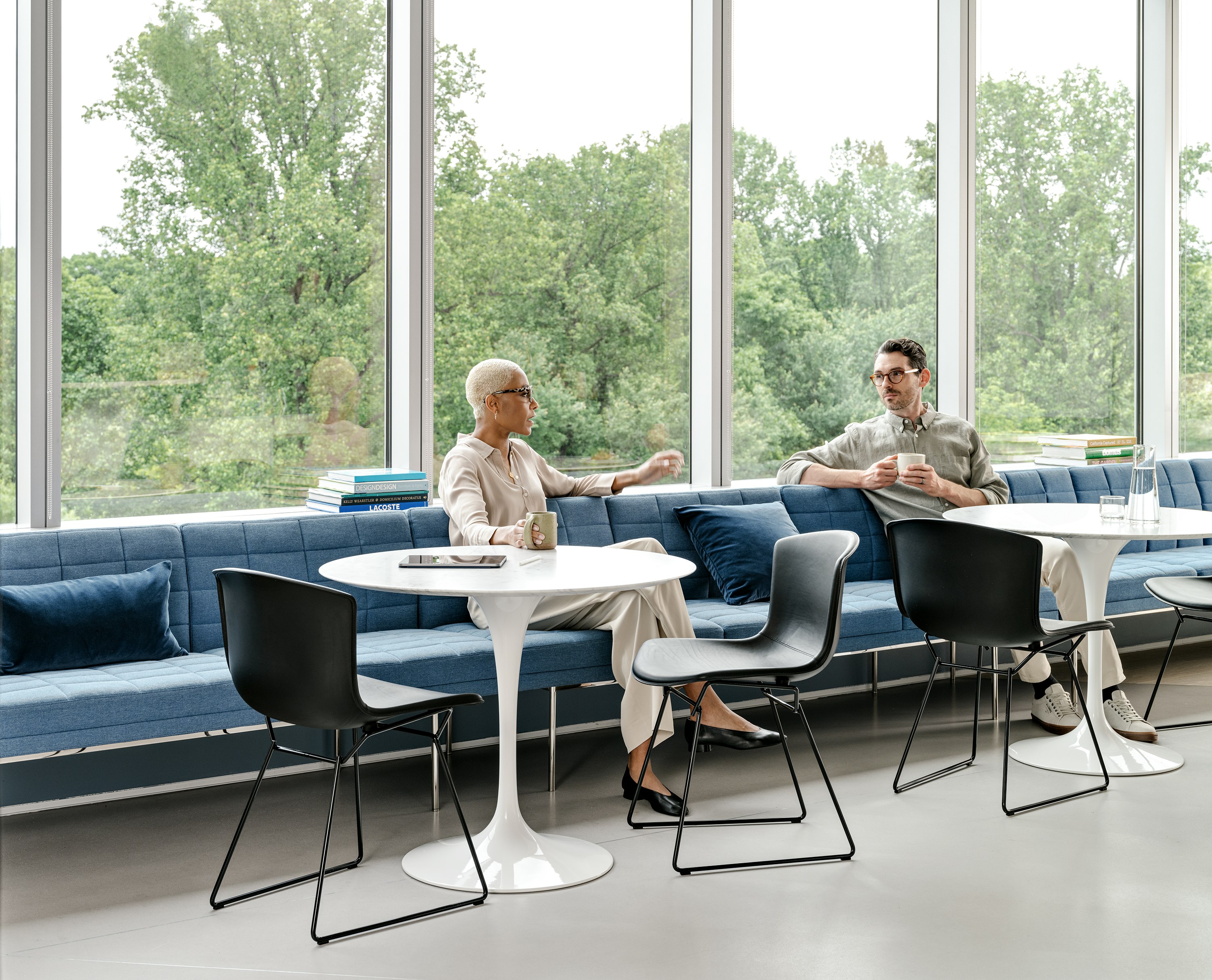 Two individuals conversing in a shared seating area furnished with Saarinen Tables, Knoll Bertoia chairs, and blue bench seating